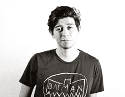 A black and white portrait of a young man with dark, curly hair. He wears a dark t-shirt with a white graphic of a bat-shaped outline and the word "BATMAN" printed on it. The background is plain white.