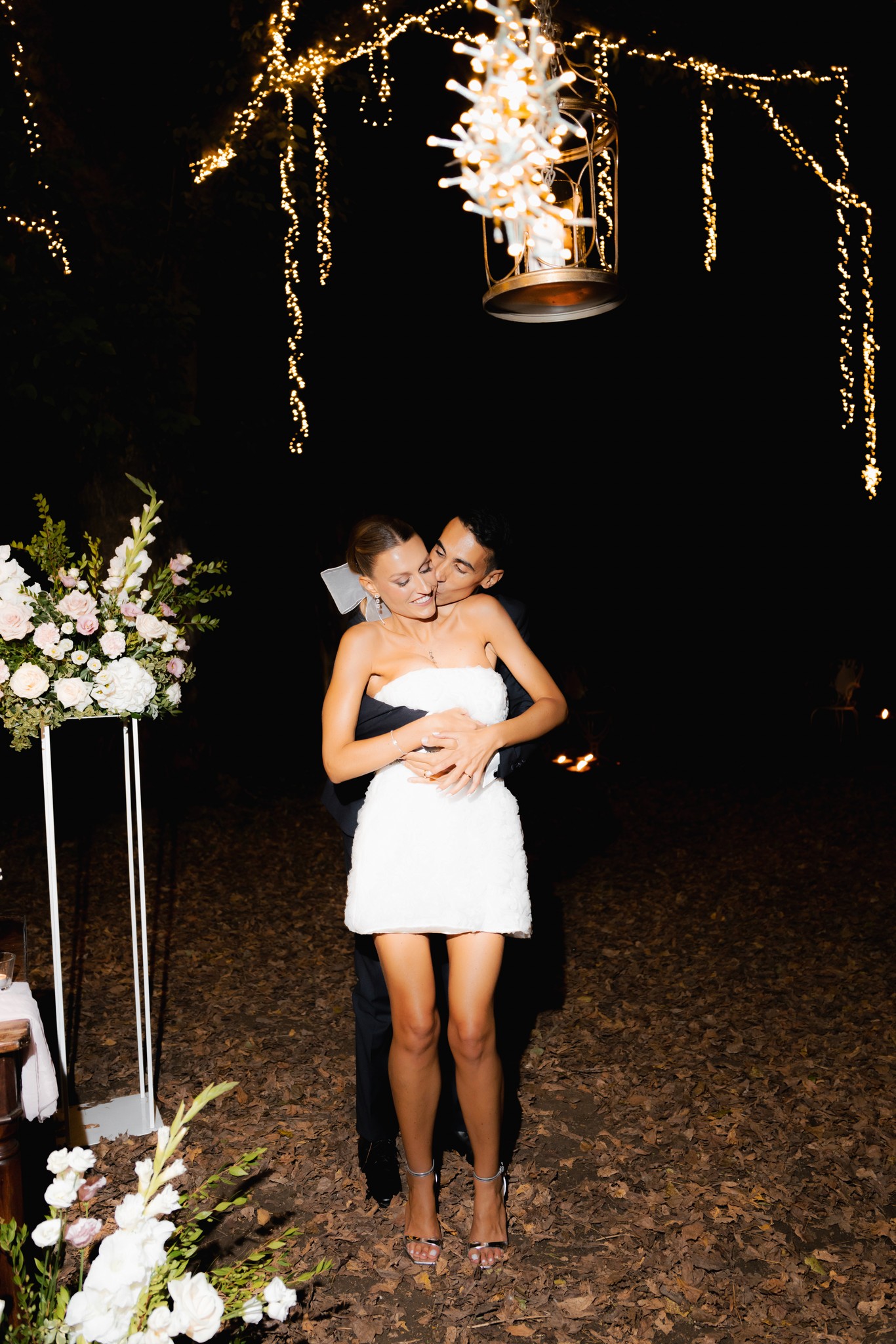 Sposa in abito bianco abbraccia lo sposo sotto decorazioni floreali dorate di sera a Villa Ferrari