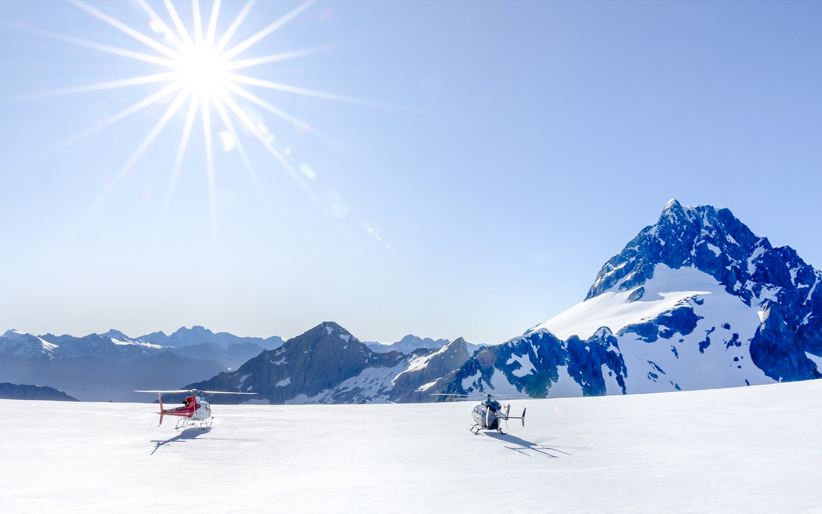 To helikoptre på en snødekt isbre i Sør-Alpene, Queenstown, under en strålende sol.