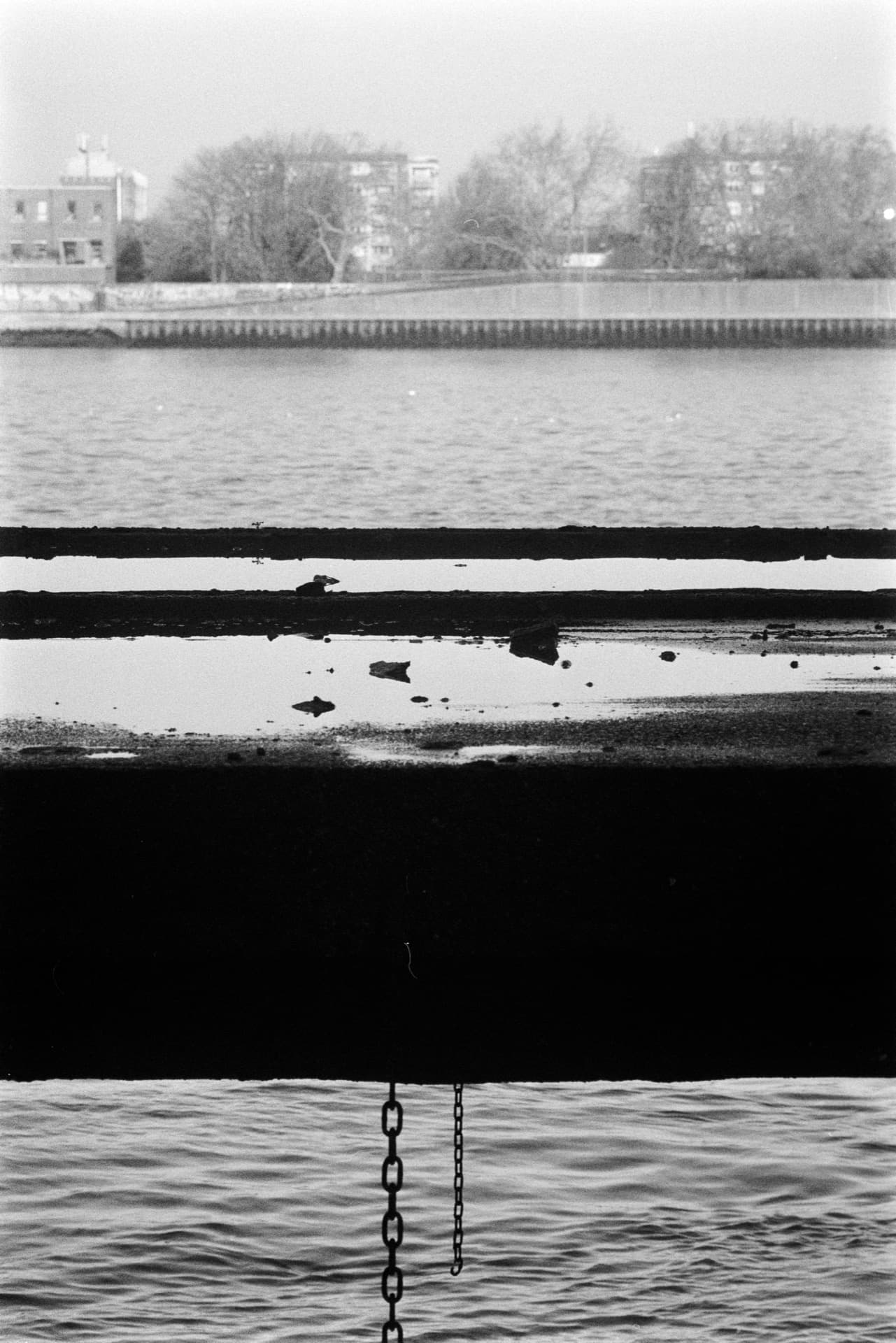 Layered Thames view with rippling water in foreground, weathered concrete pier edge with water pools, distant flood defense wall and residential towers through winter haze