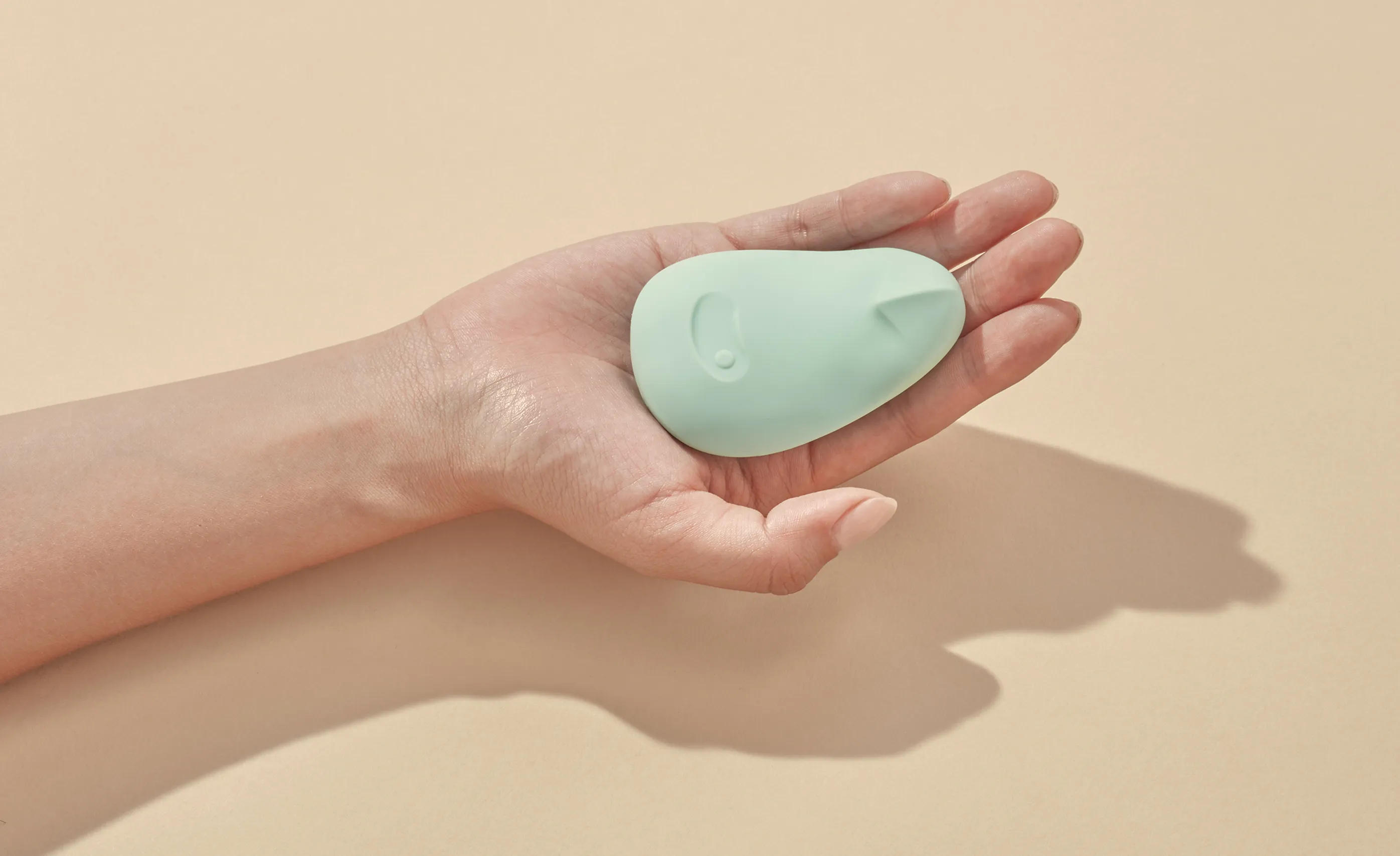 Hand holding a mint-green Dame Pom sexual wellness device on a beige background