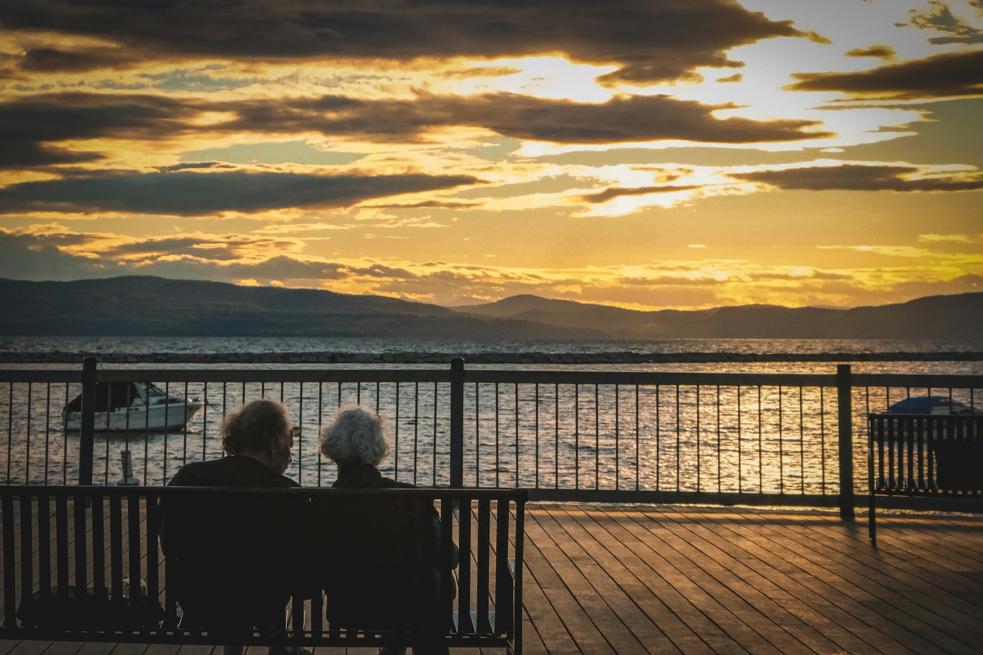 Two people sitting on a bench on a waterfront deck, watching the sunset over the water and hills.