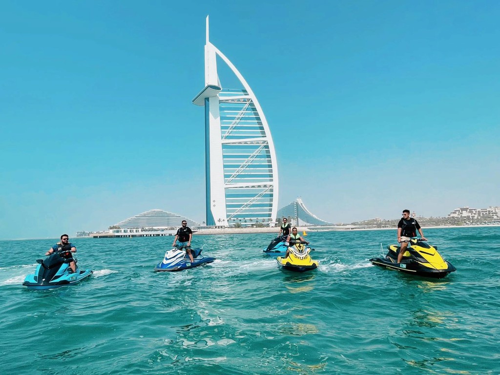 Group of riders on jet skis heading out to sea with Dubai skyline and Burj Al Arab in the background.