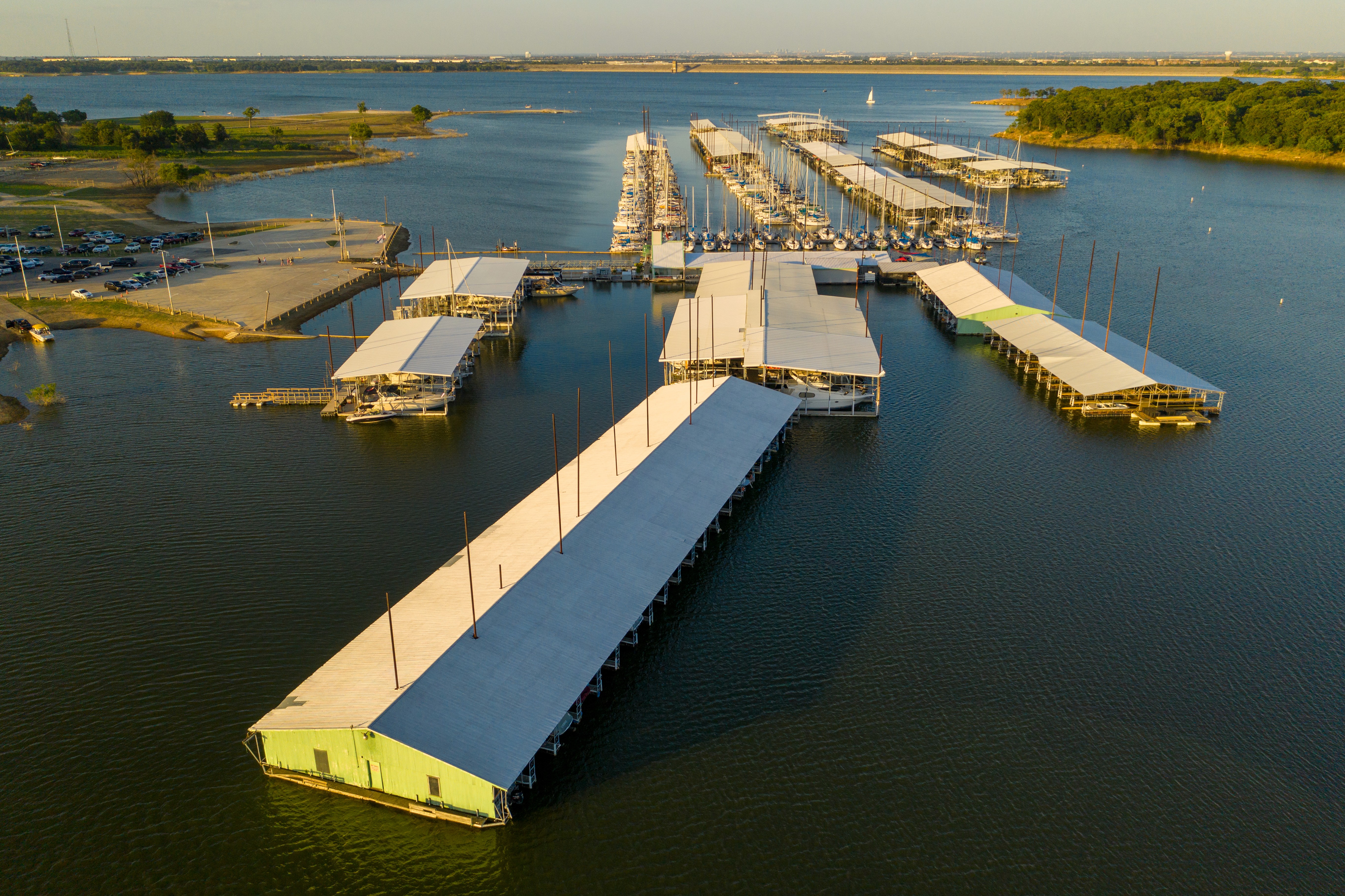 Aerial view of a marina with multiple docked boats and covered berths extending into a large calm body of water, surrounded by lush green landscape and a distant horizon under a clear blue sky.
