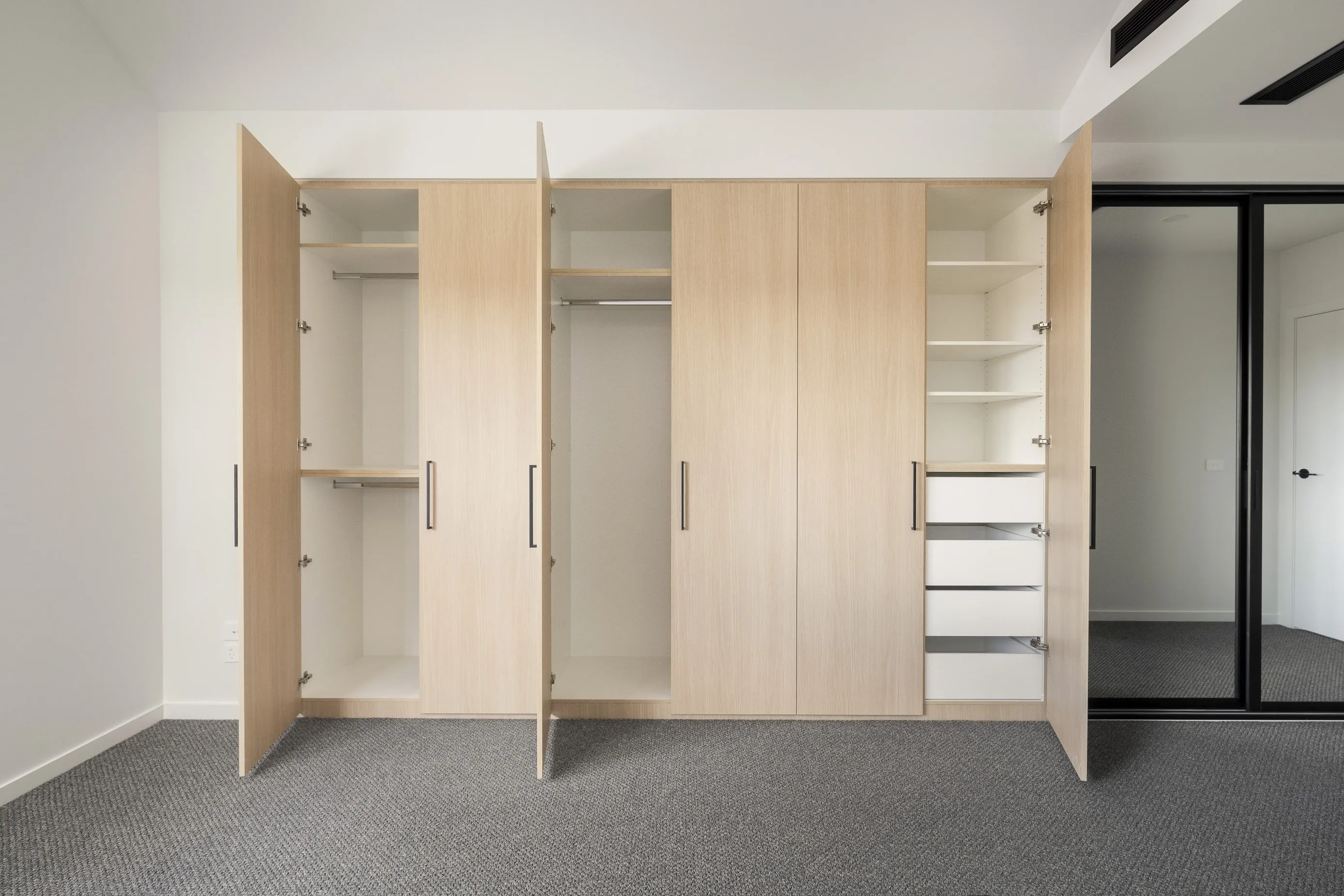 Bedroom with built-in pale-oak wardrobes opened to reveal hanging rails, shelves and drawers, grey carpet and a black-framed glazed wall to a corridor