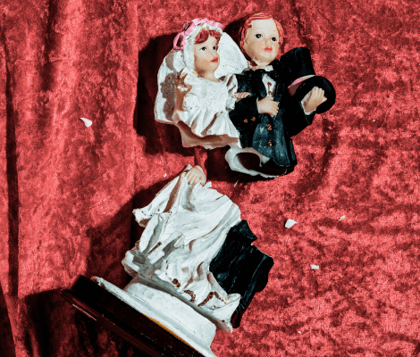 A broken wedding cake topper featuring a bride and groom figurine, with the bride's head detached from her body. The figurines are set against a rich red velvet background, highlighting the damage. The scene conveys a sense of nostalgia and loss.