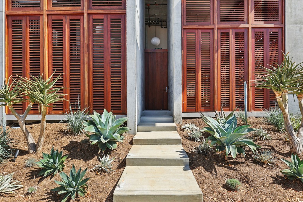 A concrete path leads to a wooden door flanked by large red shutters. Succulent plants and dry soil in the foreground create a modern, arid landscape.