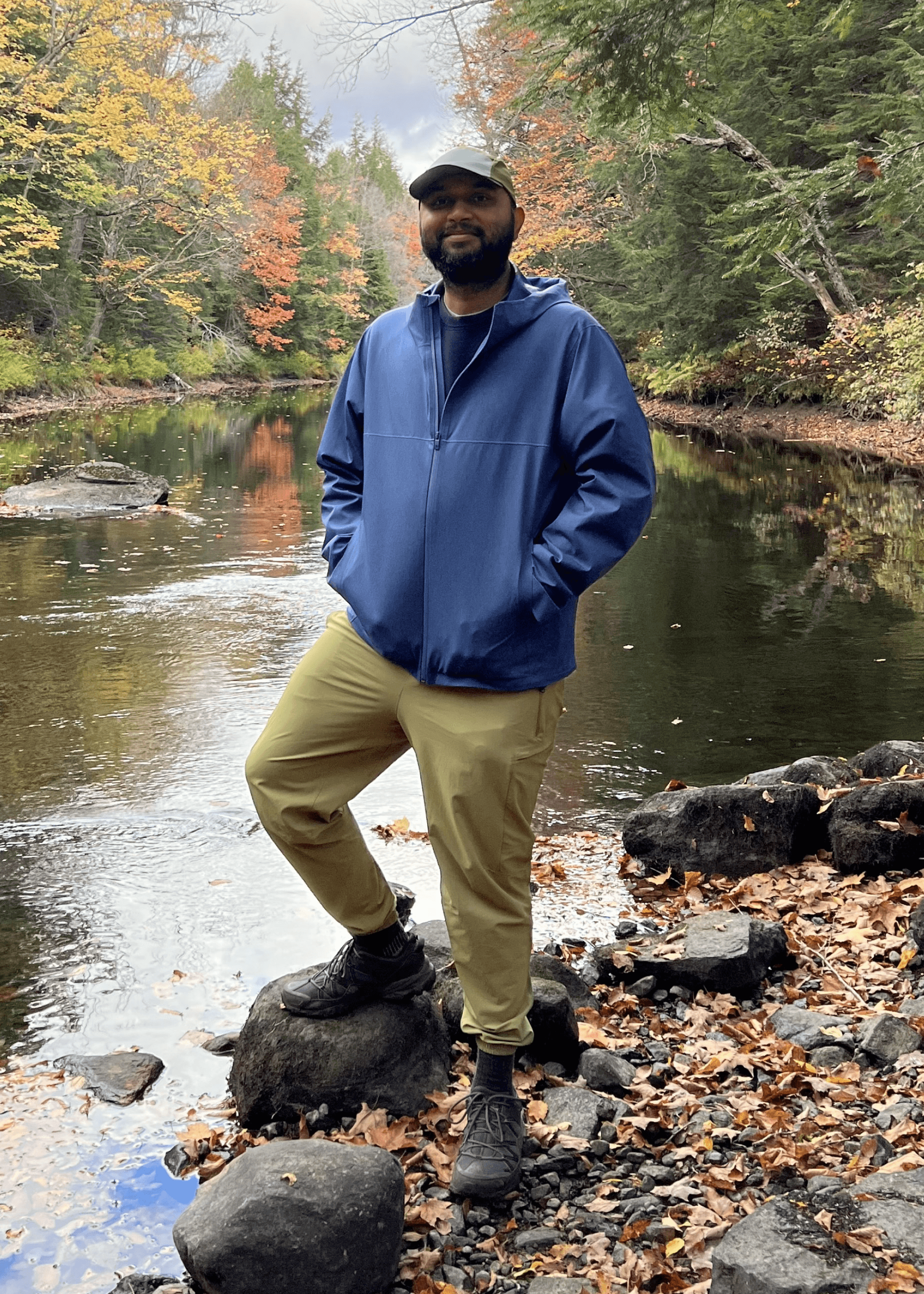 A person standing next to a stream with a backdrop of trees with fall foliage
