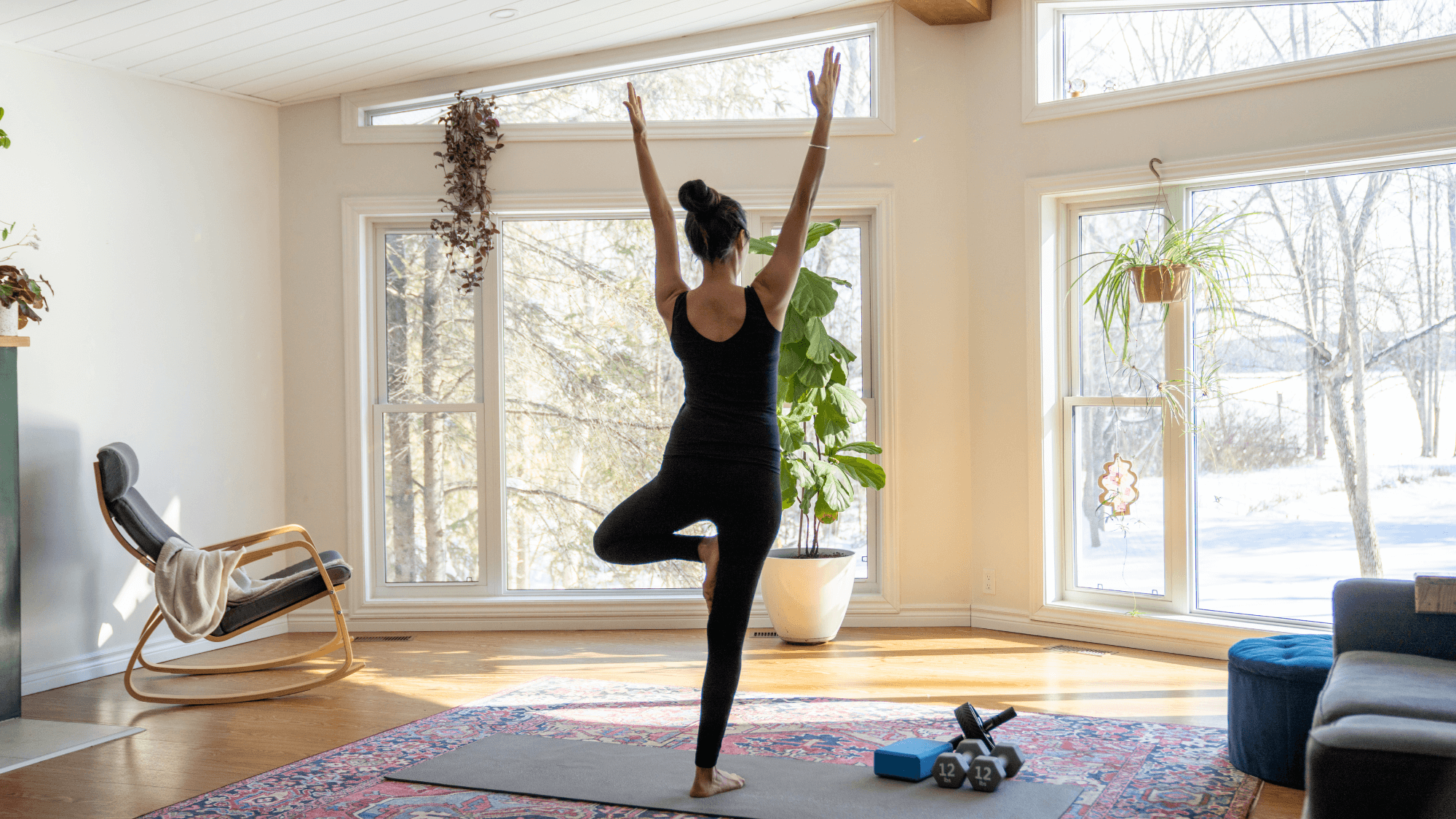 Woman practicing Tree Pose (Vrksasana) in a sunlit home yoga space, wearing black activewear, with yoga props, dumbbells, and houseplants visible against snowy winter backdrop through large windows