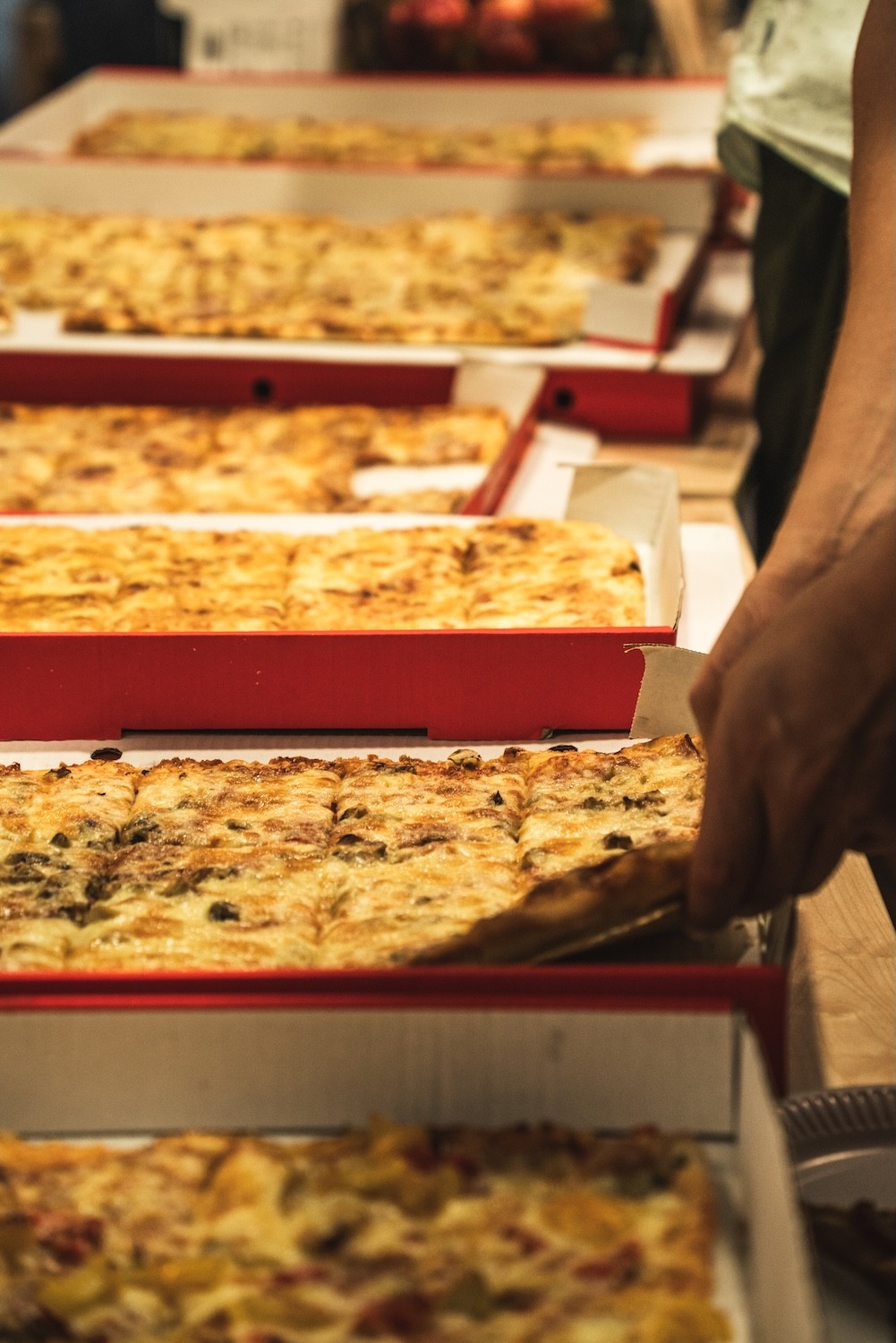 Mehrere rechteckige Pizzastücke in offenen Pizzakartons, eine Hand nimmt ein Stück heraus