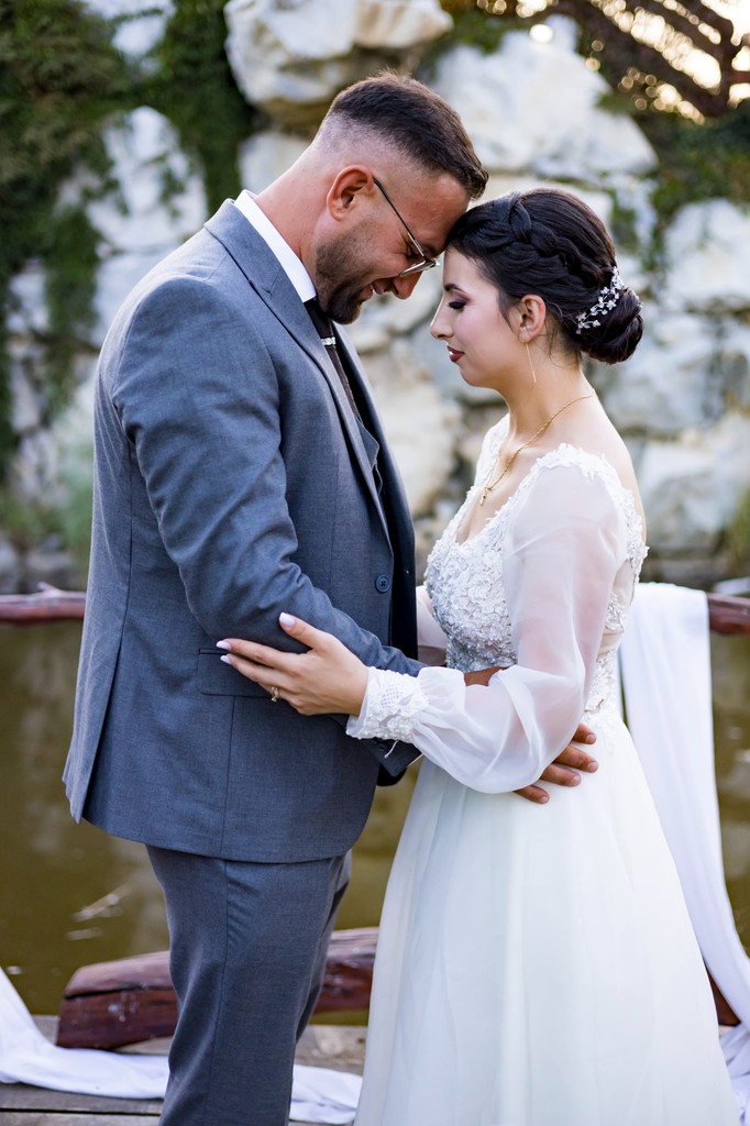 A couple in wedding attire shares a romantic kiss on a leafy path by a tranquil river.