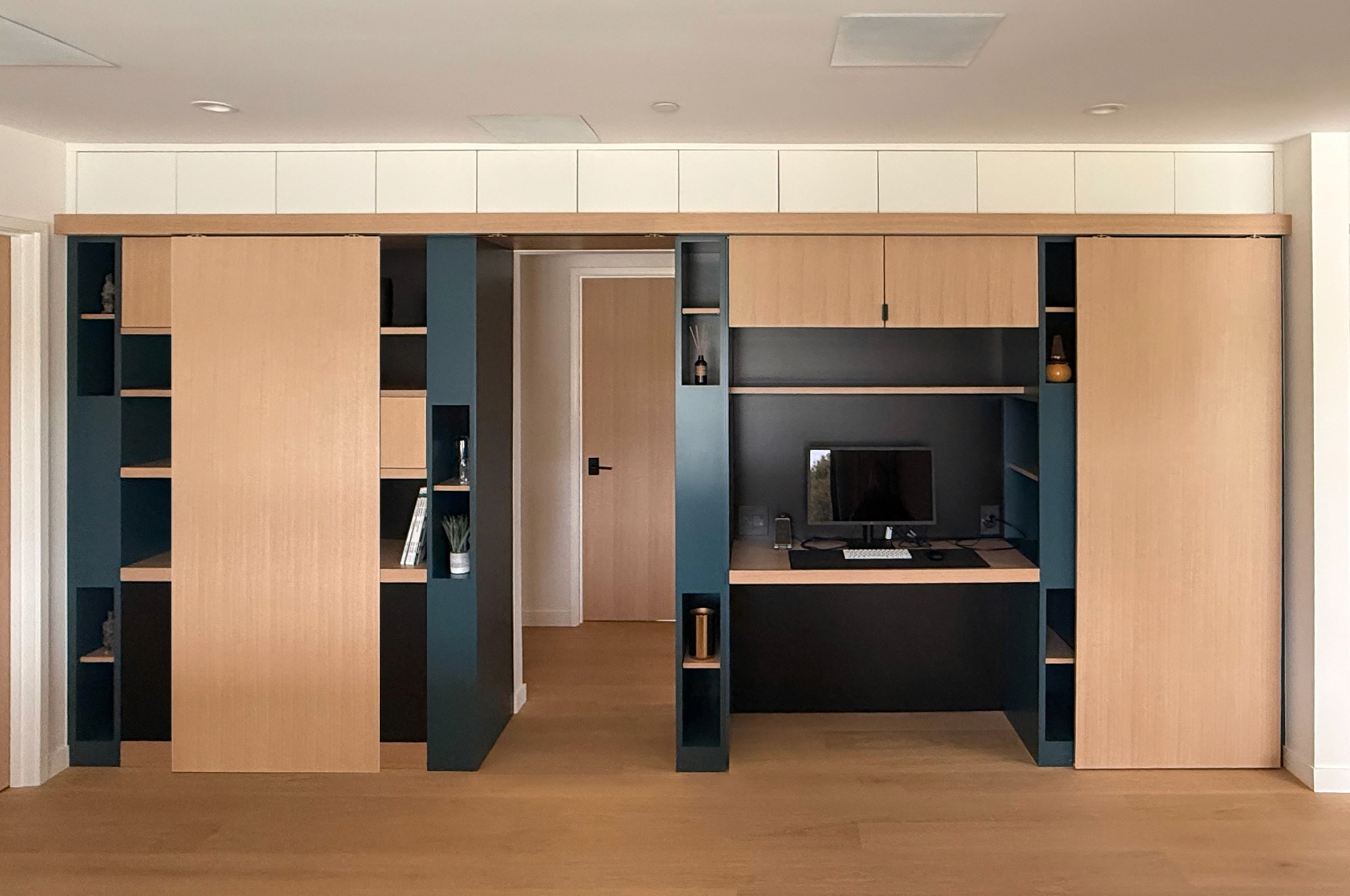 Wall of oak sliding panels and teal built-in shelving framing a hallway and a hidden desk niche