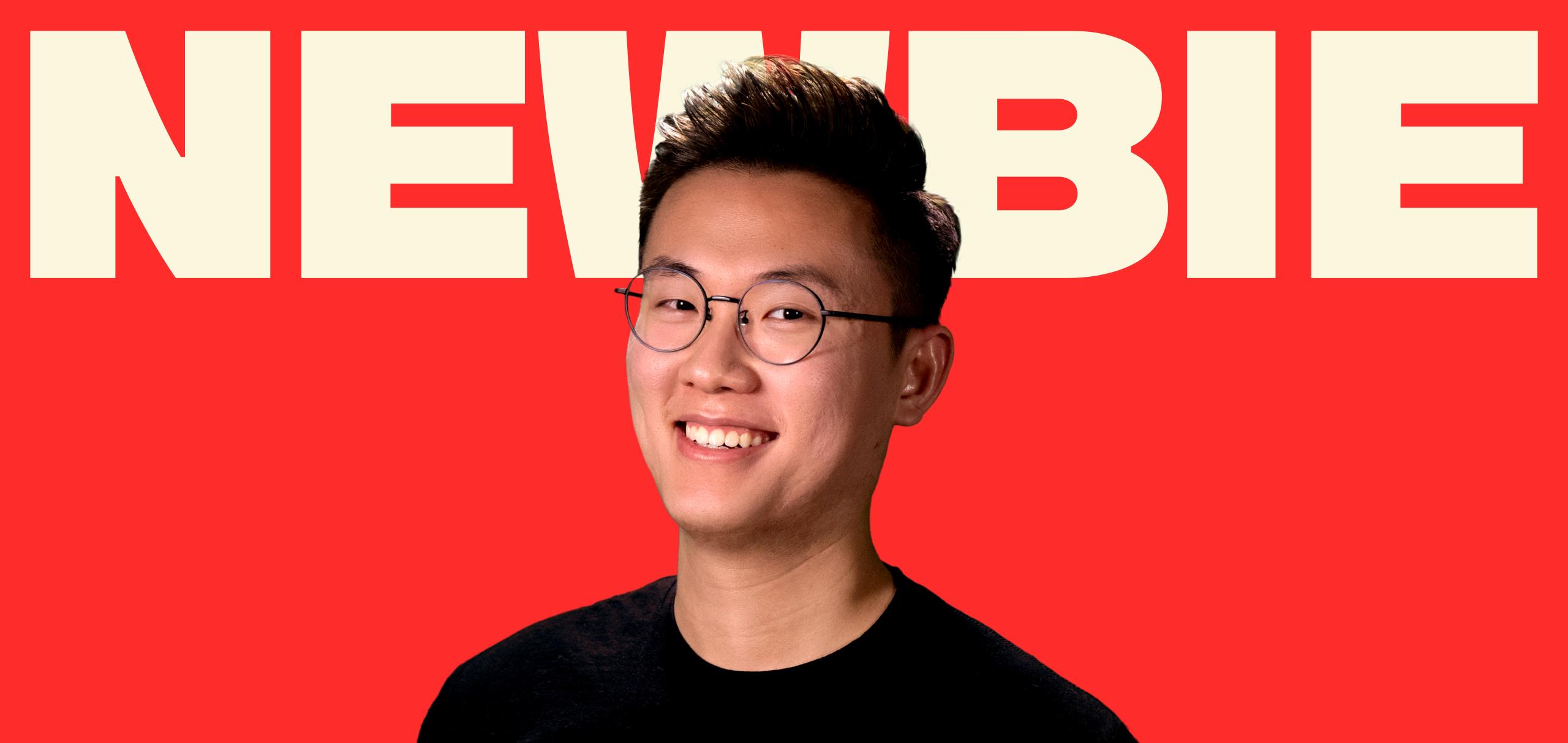 A young man with short dark hair and round glasses wears a black shirt against a red background. The word “NEWBIE” is displayed in large, off-white letters behind him.