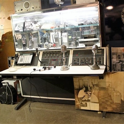 A vintage control console with microphones, buttons, and switches, flanked by communication equipment, and a black-and-white photo on the floor.