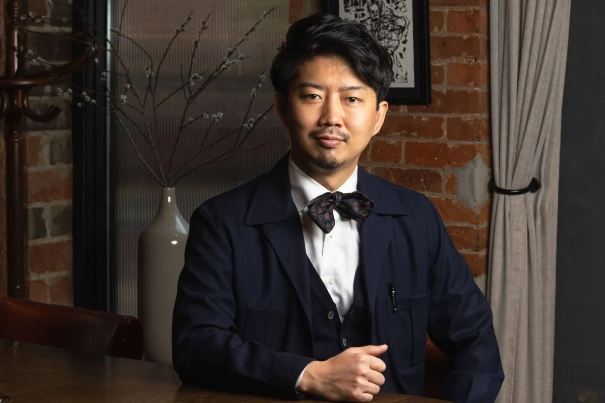 Bartender Takuma Watanabe in a navy suit and bow tie, seated against a brick wall