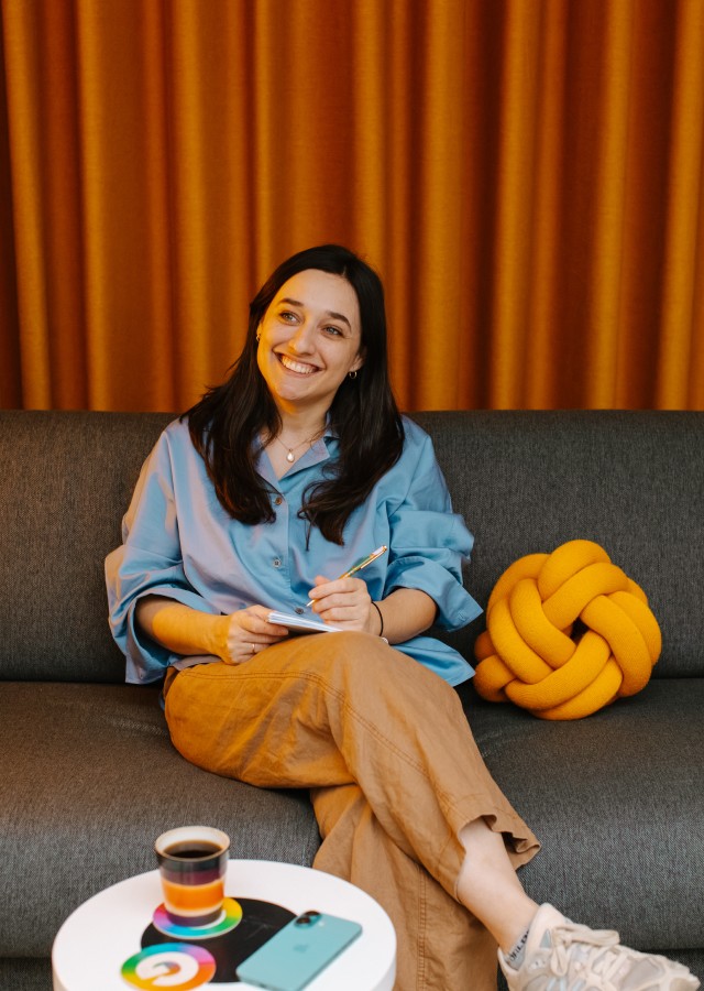 Woman smiling on a sofa holding a notepad, with a coffee and yellow knot cushion nearby.