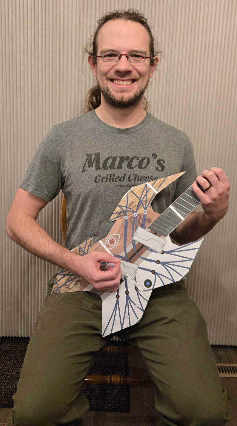 Cardboard mockup of Genesis custom multiscale guitar resting on leg in seated playing position