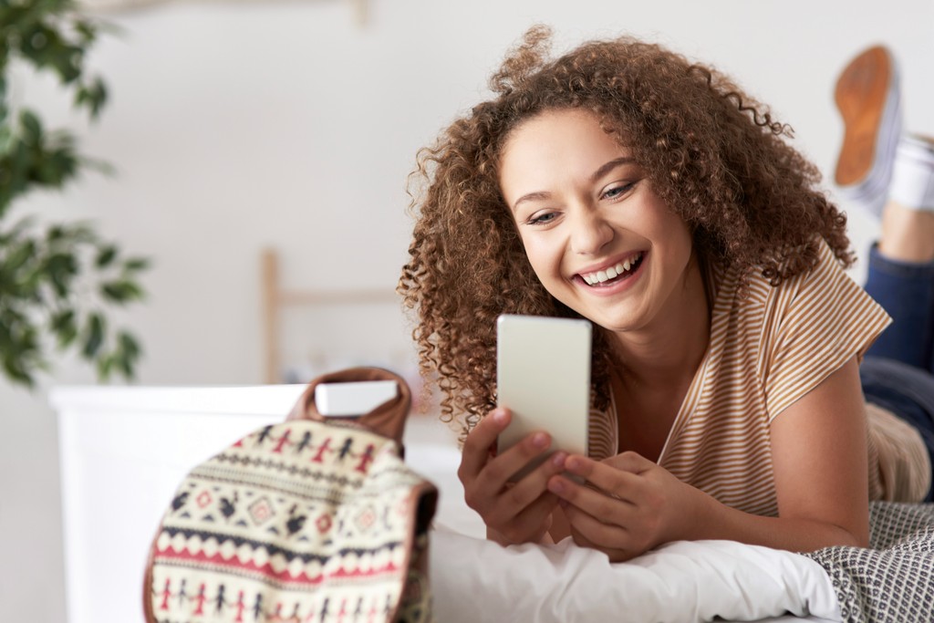 A cheerful young woman with curly hair lies on her bed, smiling and looking at her smartphone, capturing a moment of happiness and digital connectivity in a cozy, casual setting.