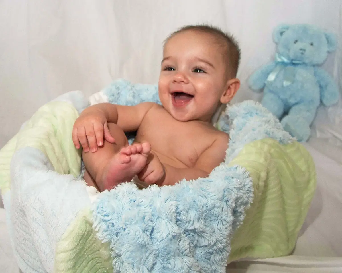A cheerful baby laughing while sitting in a cozy basket lined with soft blue and green blankets, with a light blue teddy bear in the background, captures a heartwarming scene in Bradenton, FL.