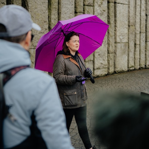 一位女士撑着紫色雨伞与一群人交谈，身后是一堵石墙。部分人群出现在前景中。