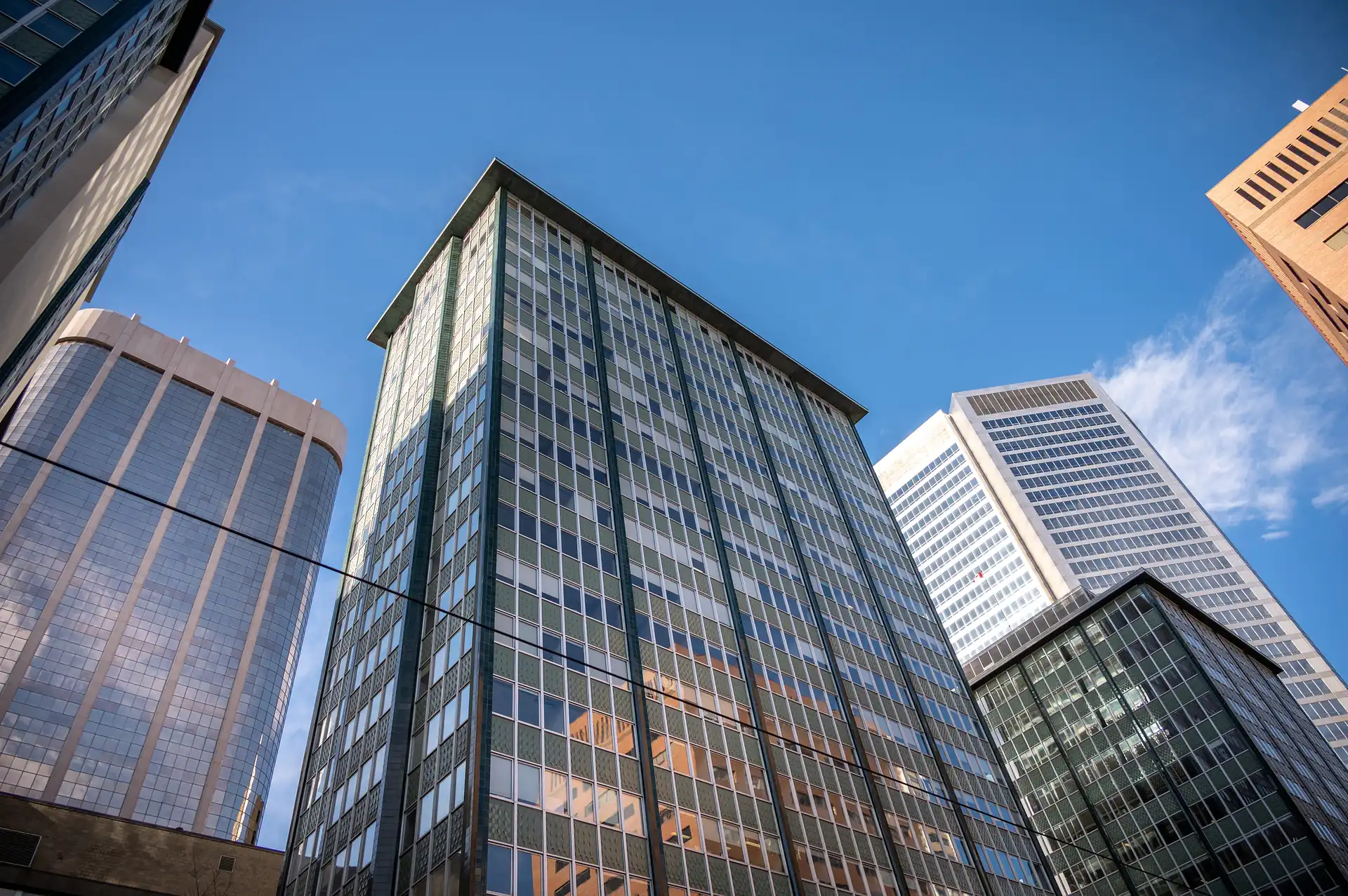 A low-angle view of several commercial skyscrapers set against a clear blue sky. Standing out in the center is a tall building with a gray and dark-green grid-patterned façade, its windows reflecting the surrounding structures.