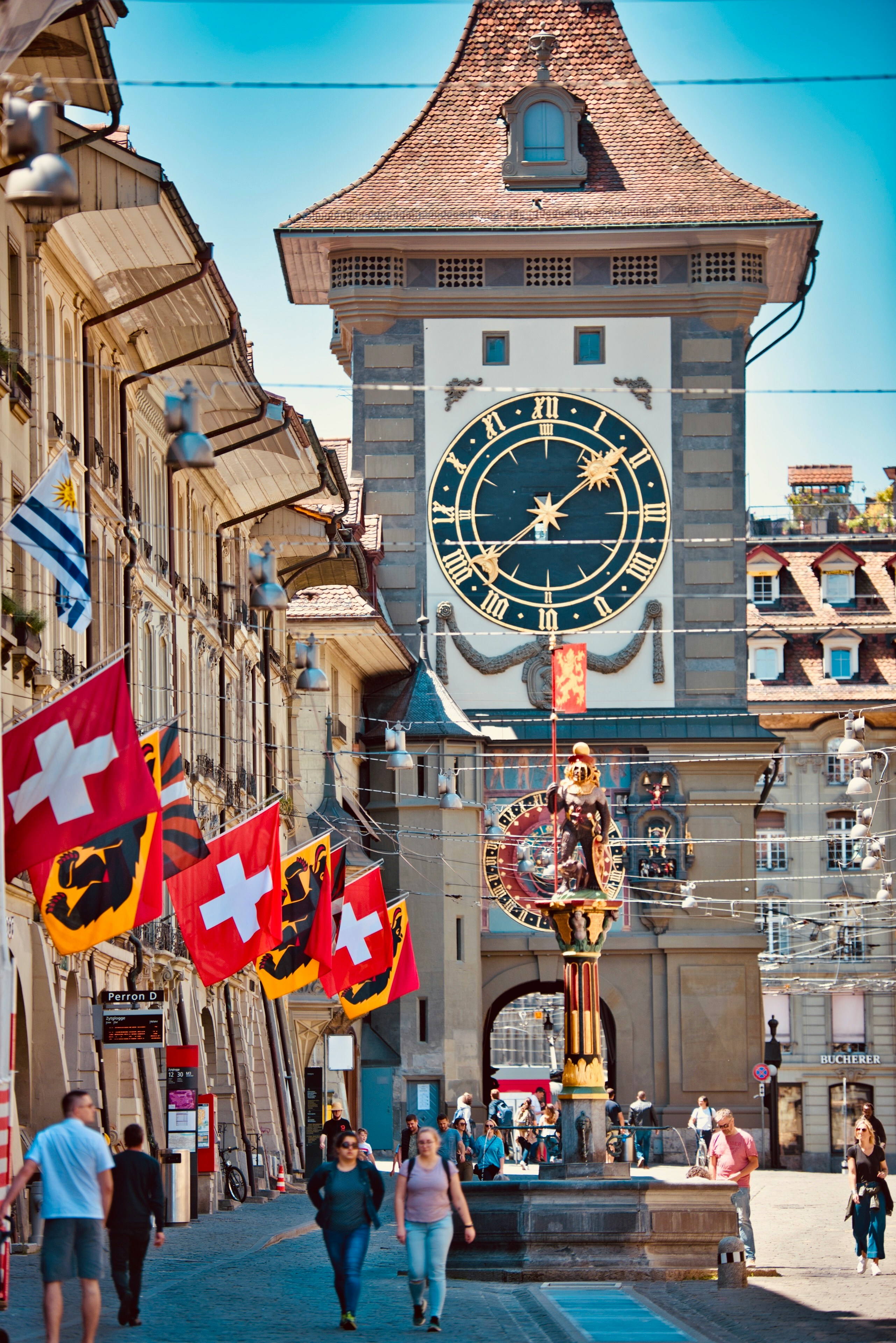 Bern’s Zytglogge clock tower with Swiss flags along the street and people walking in the historic city center.