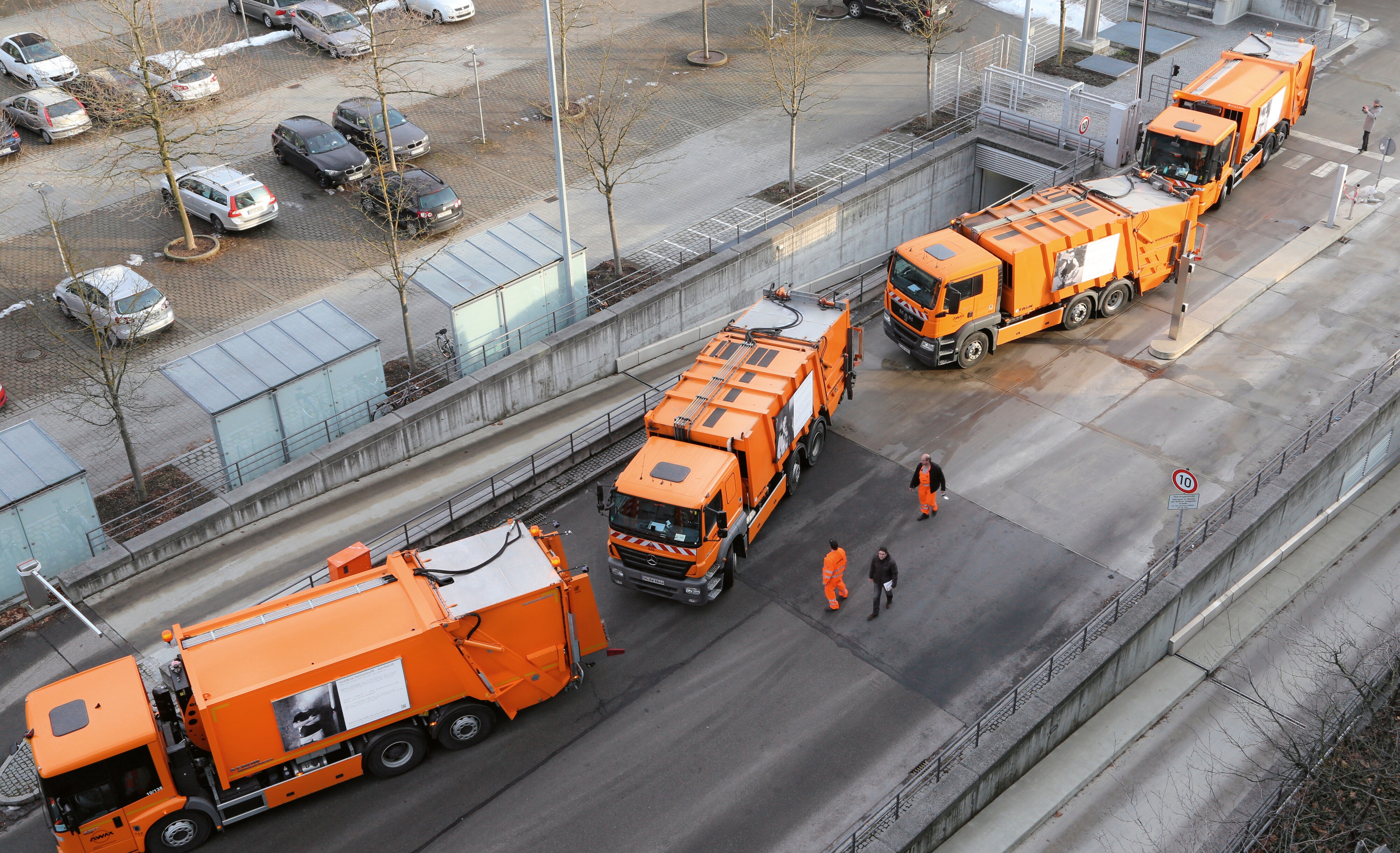 Kolonne orangefarbener Müllwagen auf einem Hof von oben, dazwischen Arbeiter in Warnkleidung