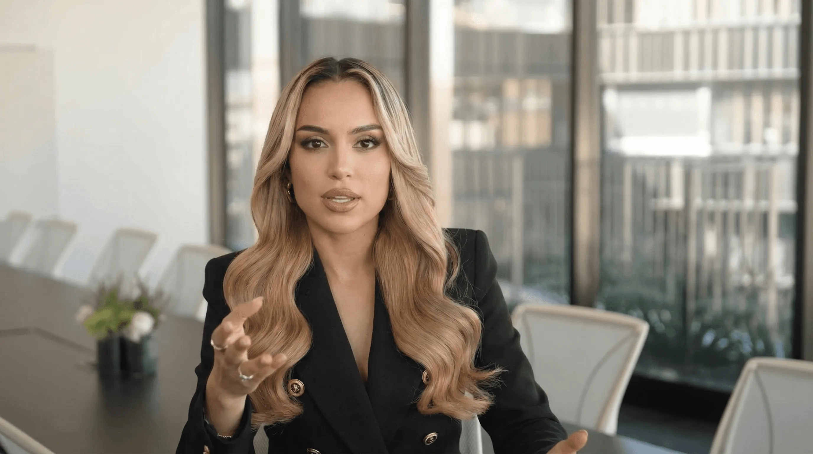 Persona hablando directamente a cámara en una sala de juntas moderna, con mesas, sillas y grandes ventanales al fondo.