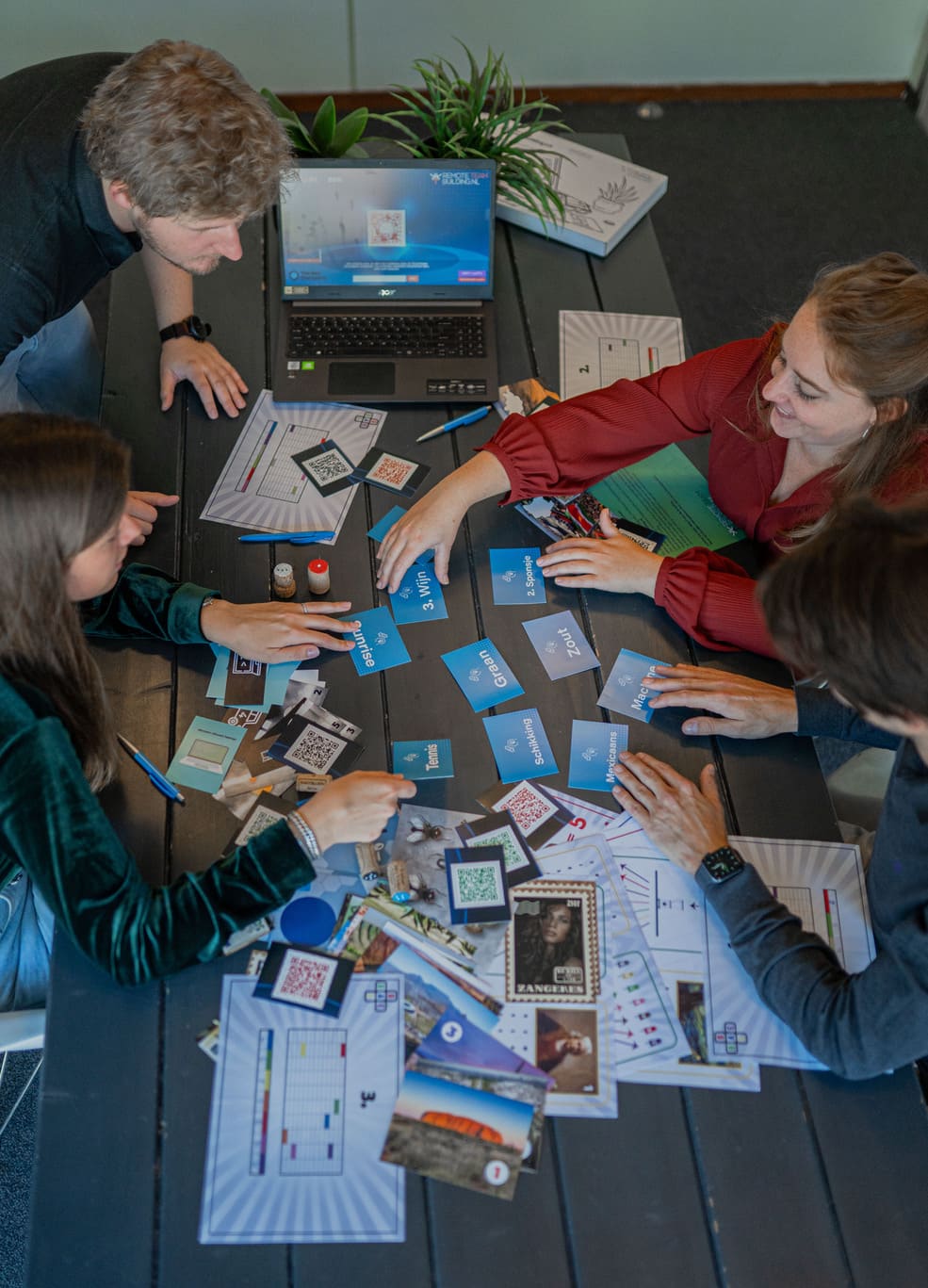 Collega’s werken samen aan een online en hybride teambuildingactiviteit, waarbij ze puzzels en opdrachten oplossen met kaarten en een laptop op tafel.