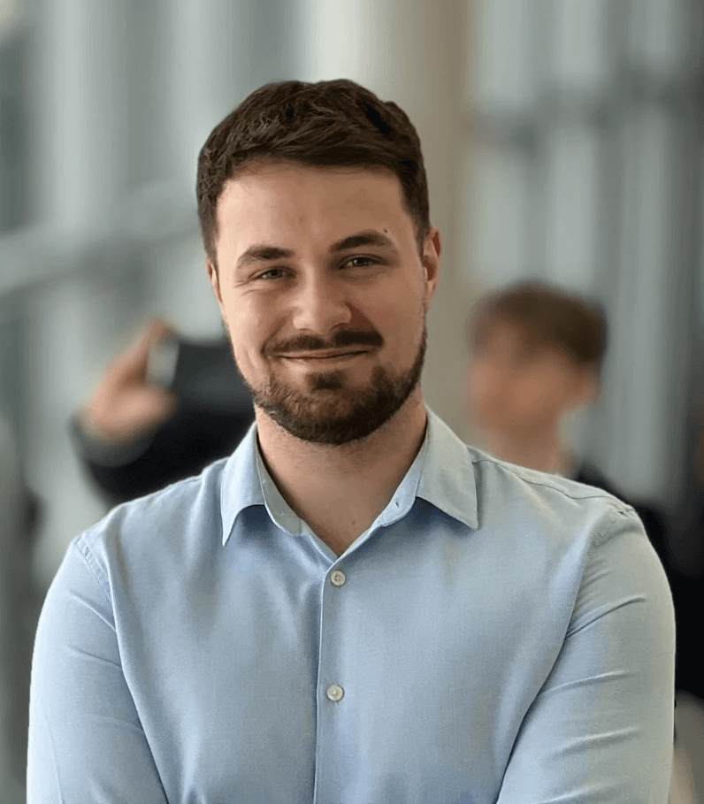 Close-up portrait of a person wearing a gray insulated hooded jacket with the hood up, showing their face framed by the hood against a light background.