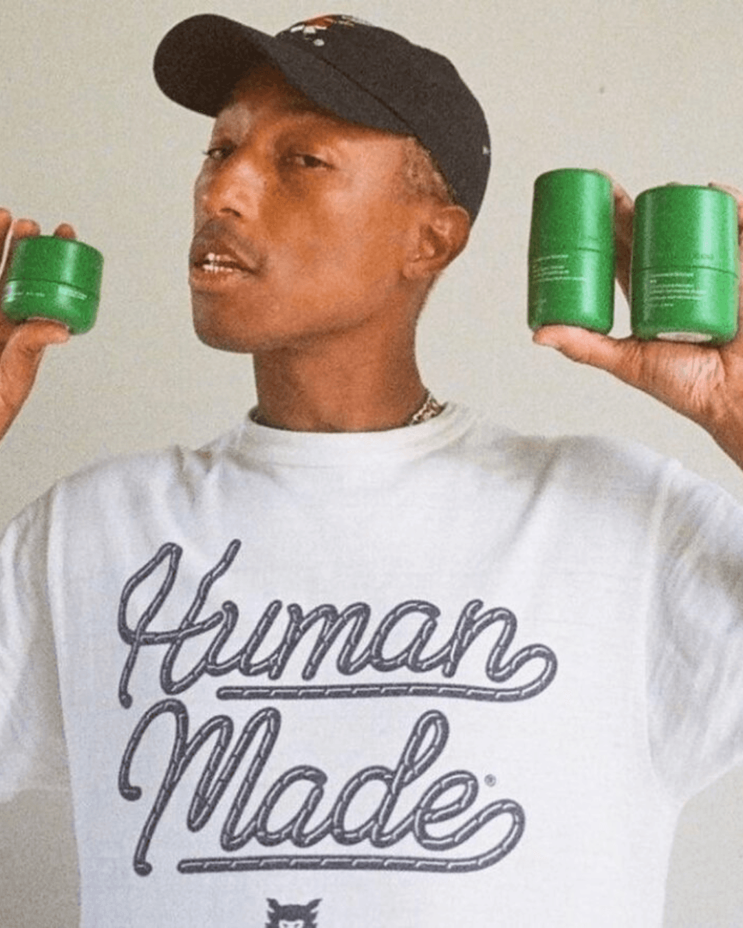 Pharrell, wearing a white "Human Made" shirt holds two green cans, posing with a thoughtful expression.