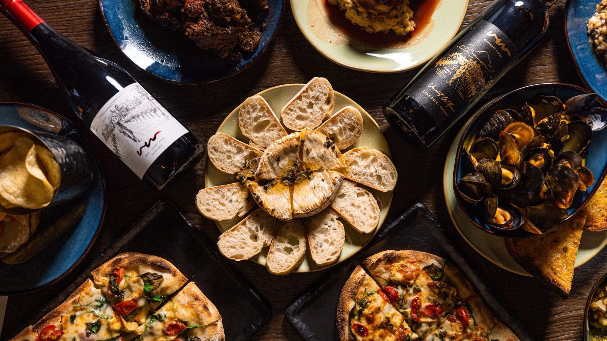 Flatbreads, baked brie, mussels and estate wine across a dark wood table