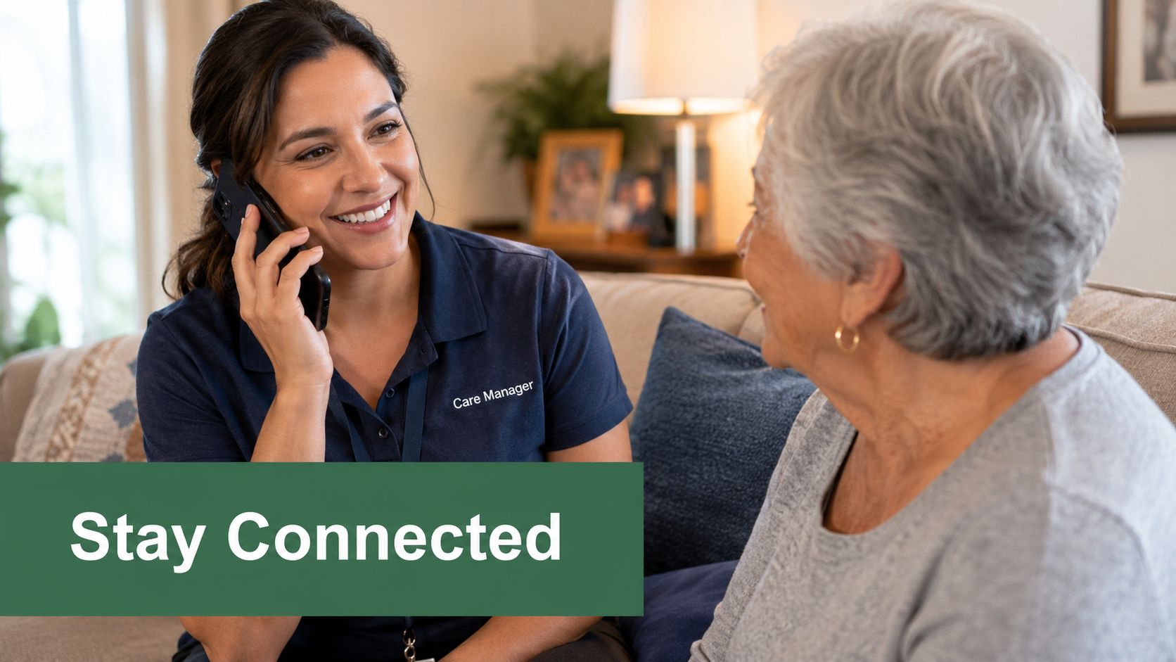 A smiling care manager speaks on the phone while sitting with an elderly woman on a couch.