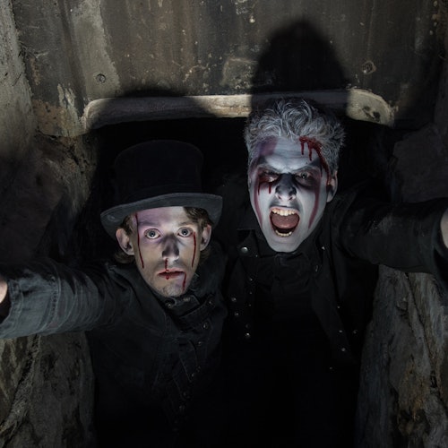 Two people with dramatic makeup and fake blood look upwards from a dark, enclosed space; one wears a top hat.