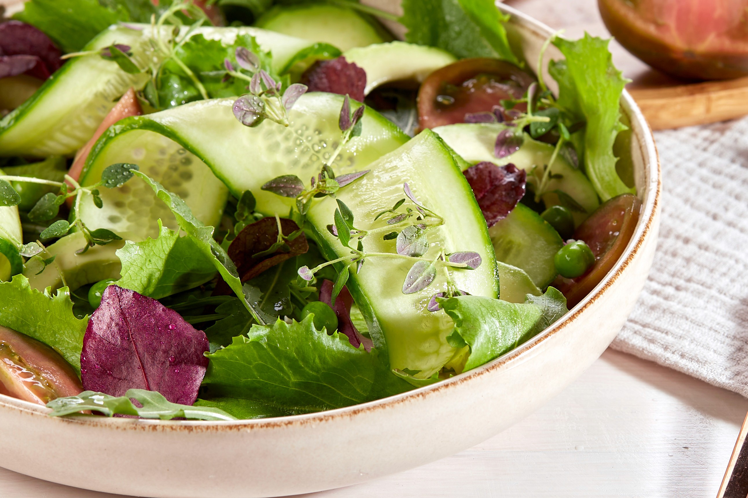 A fresh garden salad in a ceramic bowl features crisp cucumber slices, vibrant green leafy lettuce, ripe tomato wedges, and a sprinkle of delicate herbs, set on a light-colored table with a wooden spoon nearby.
