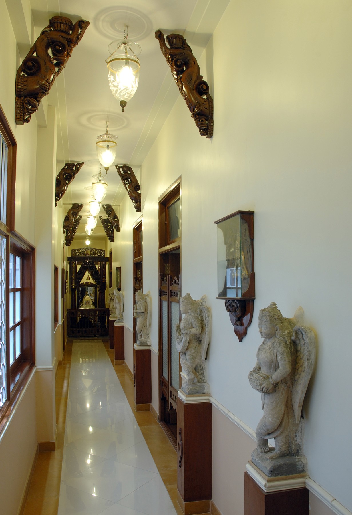 Long corridor lined with carved wooden brackets, pendant lights and sculptural angel statues in a heritage residence.