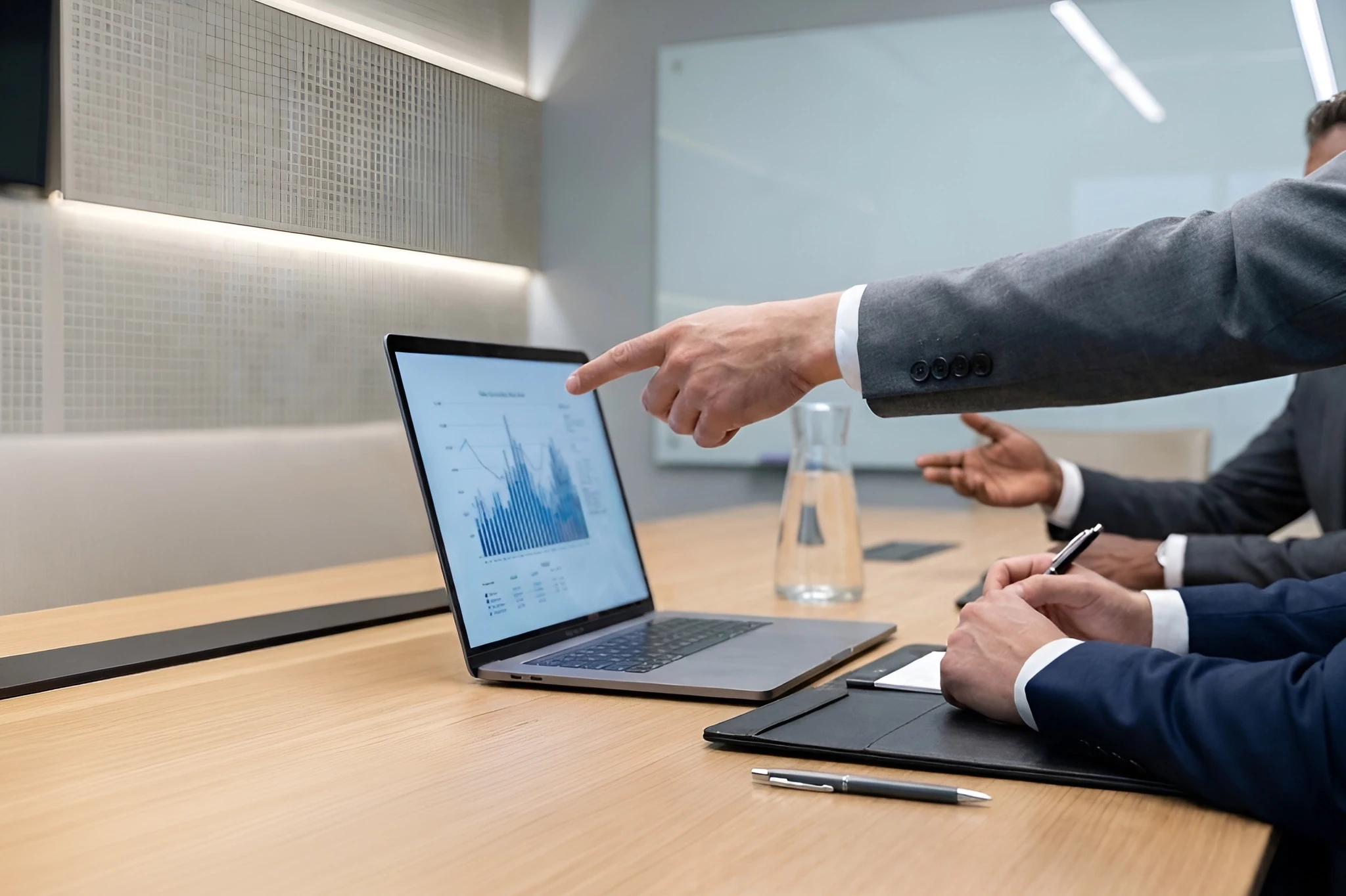Professional pointing to a line and bar graph on a laptop screen during a business meeting in a boardroom.