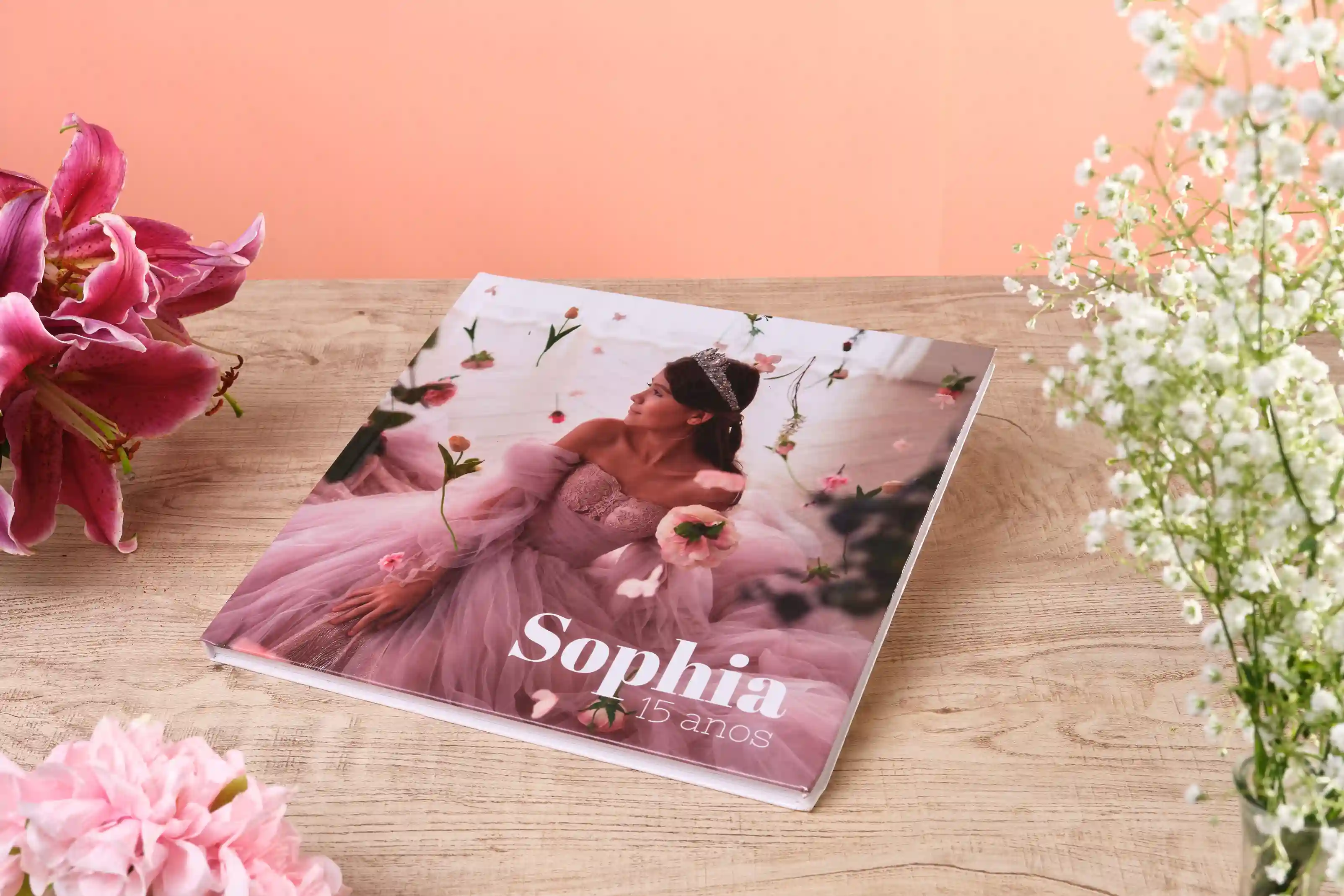 Fotolivro personalizado de 15 anos com capa da jovem Sophia vestindo vestido rosa, decorado com flores, apoiado sobre mesa de madeira com arranjos florais ao redor.