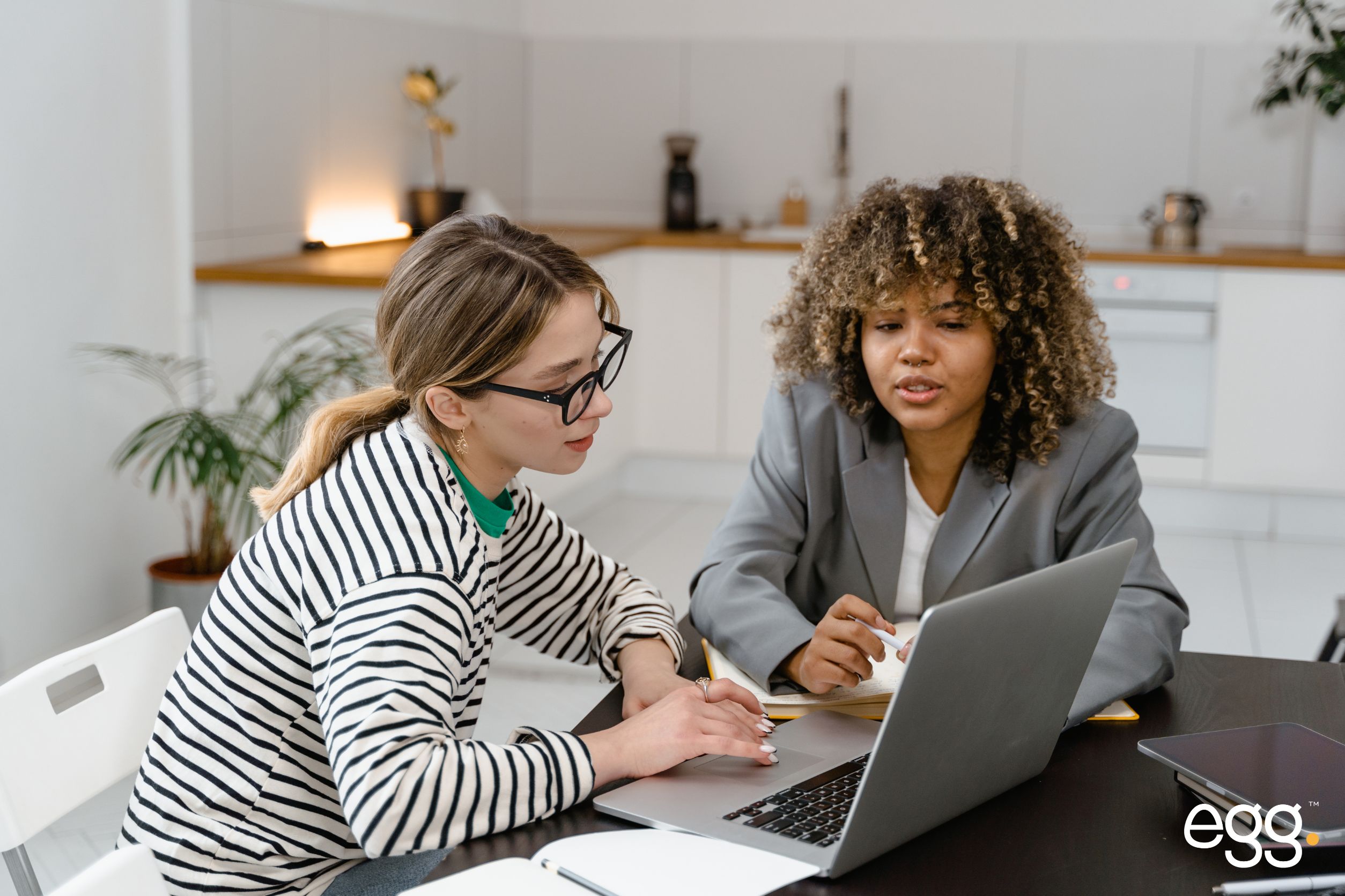 Two women collaborating in a modern office setting, with one wearing glasses and a striped sweater and the other in a blazer. They are sitting at a table, discussing and taking notes while looking at a laptop screen. A small "egg" logo is displayed in the bottom right corner.