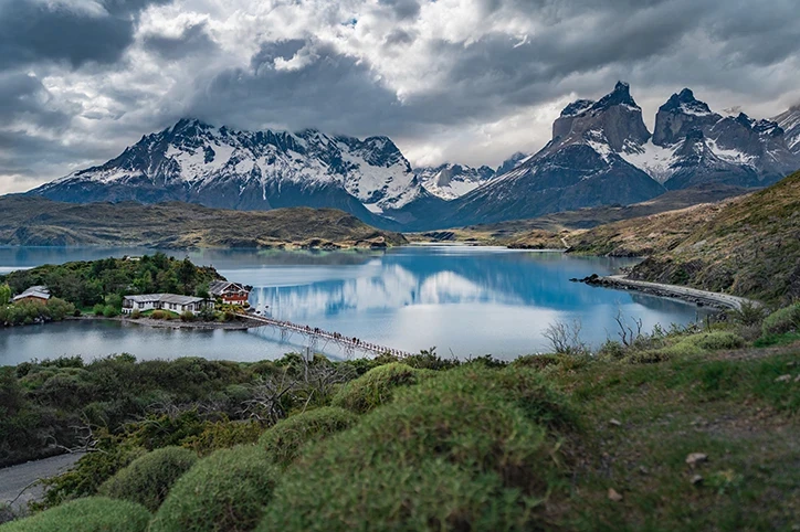 Panorama Patagonii z jeziorem, drewnianym mostem, domkami i ośnieżonymi szczytami Torres del Paine pod burzowym niebem