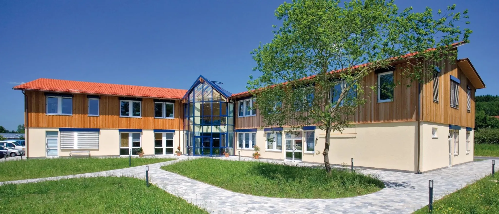 Modern Holzbau-Gebäude mit Glasfassanade, rotem Ziegeldach und grüner, gepflegter Außenanlage.