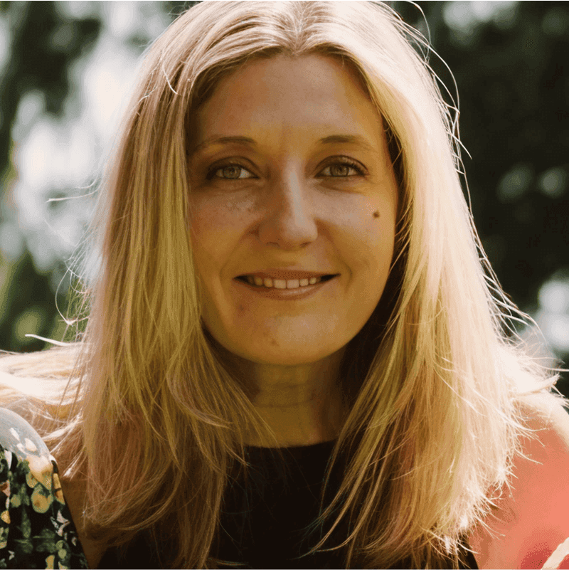 Close-up portrait of a smiling woman with long, light brown hair, surrounded by greenery in natural light.