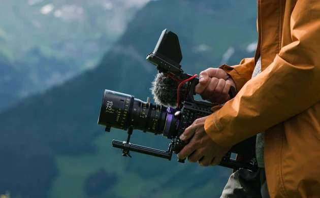 A filmmaker holding a cinema camera with a DZOFILM Arles zoom lens against a mountain backdrop