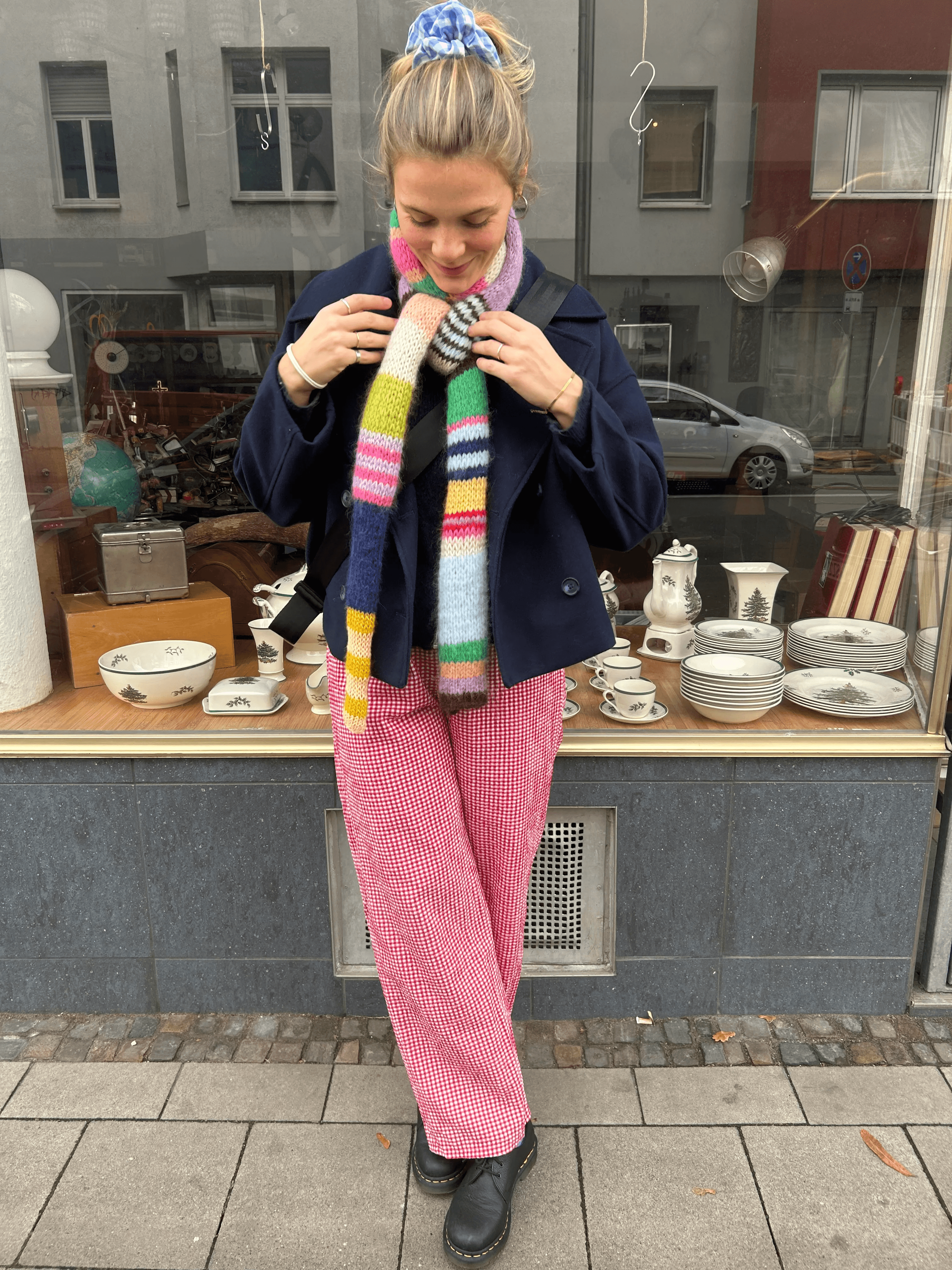 A woman stands in front of a shop window displaying vintage crockery, wearing a colorful striped scarf, a navy jacket, pink patterned pants, and black boots.