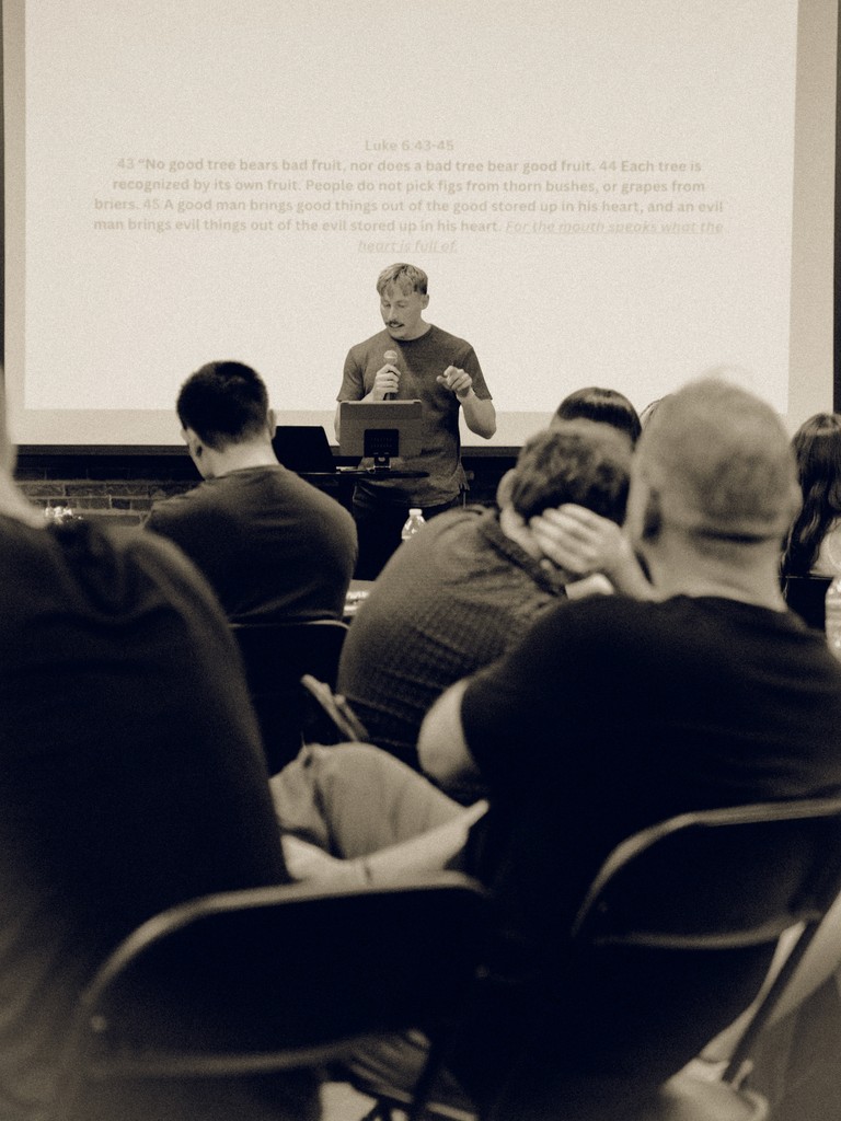 Room full of people at roundtables conversating, connecting, and sharing stories. Speaker standing in front of room preaching.