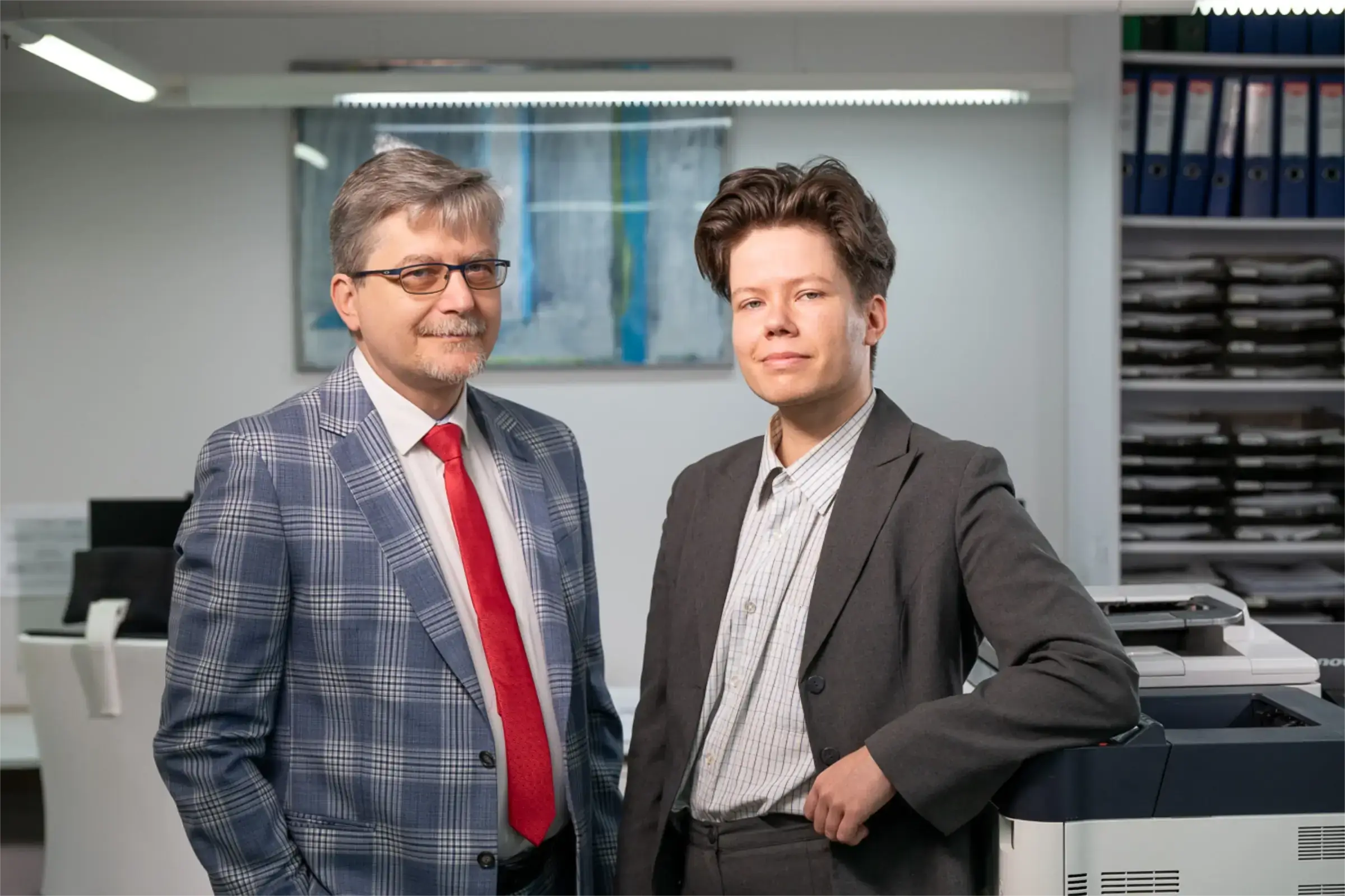 Dwóch mężczyzn w garniturach, starszy z okularami i czerwonym krawatem, młodszy uśmiechnięty, na tle biurowym.