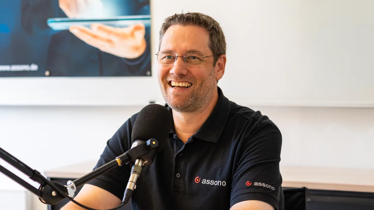 Thomas Bahn sitzt lächelnd vor einem Mikrofon und trägt ein schwarzes Polo-Shirt mit assono-Logo. Im Hintergrund ist ein heller Büroraum zu sehen.