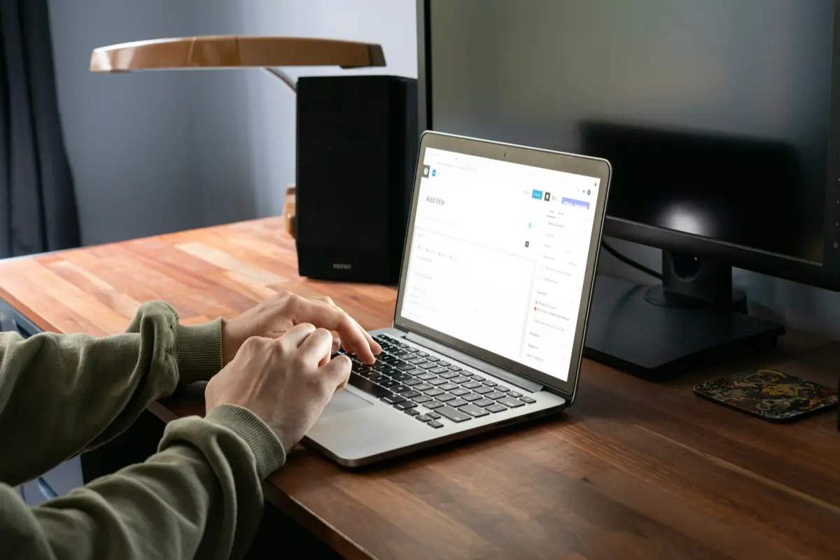 A person typing on a laptop at a wooden desk, with a desk lamp and a speaker in the background.