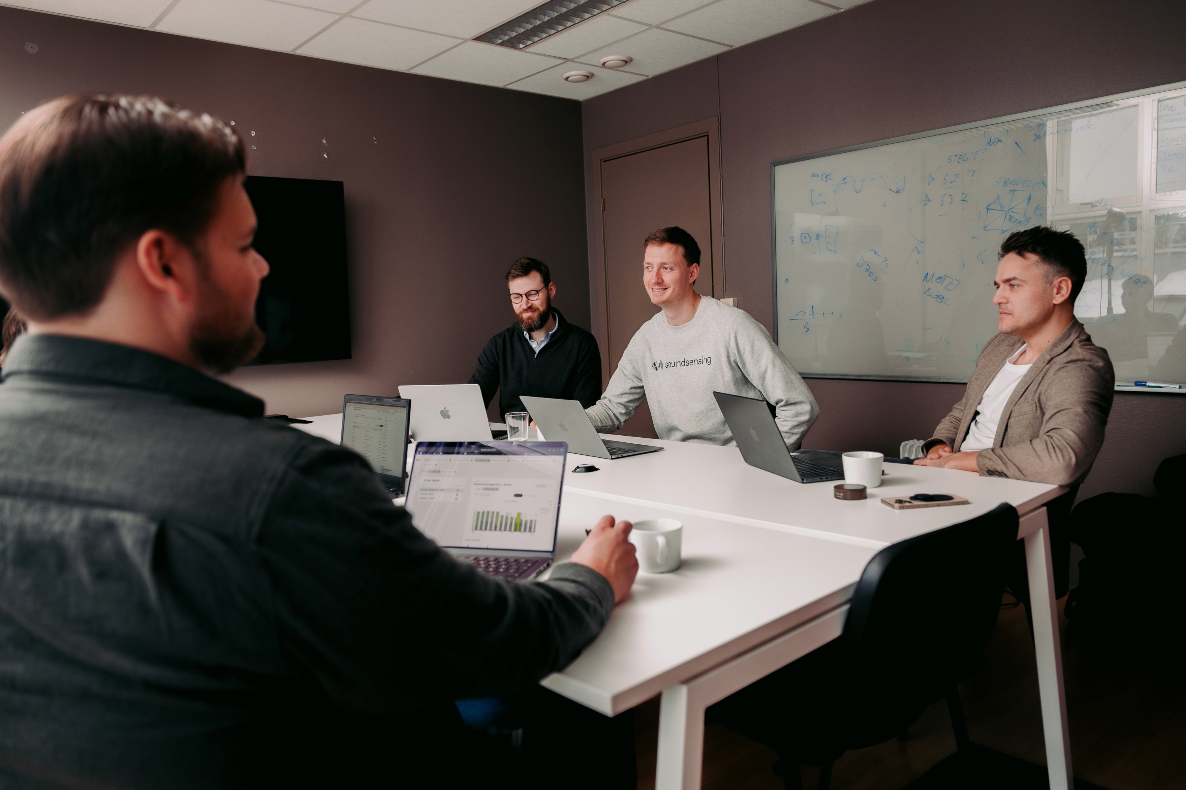 A group of people seated around a conference table in an office setting, engaged in a professional meeting with laptops, notebooks, and coffee cups scattered on the table.