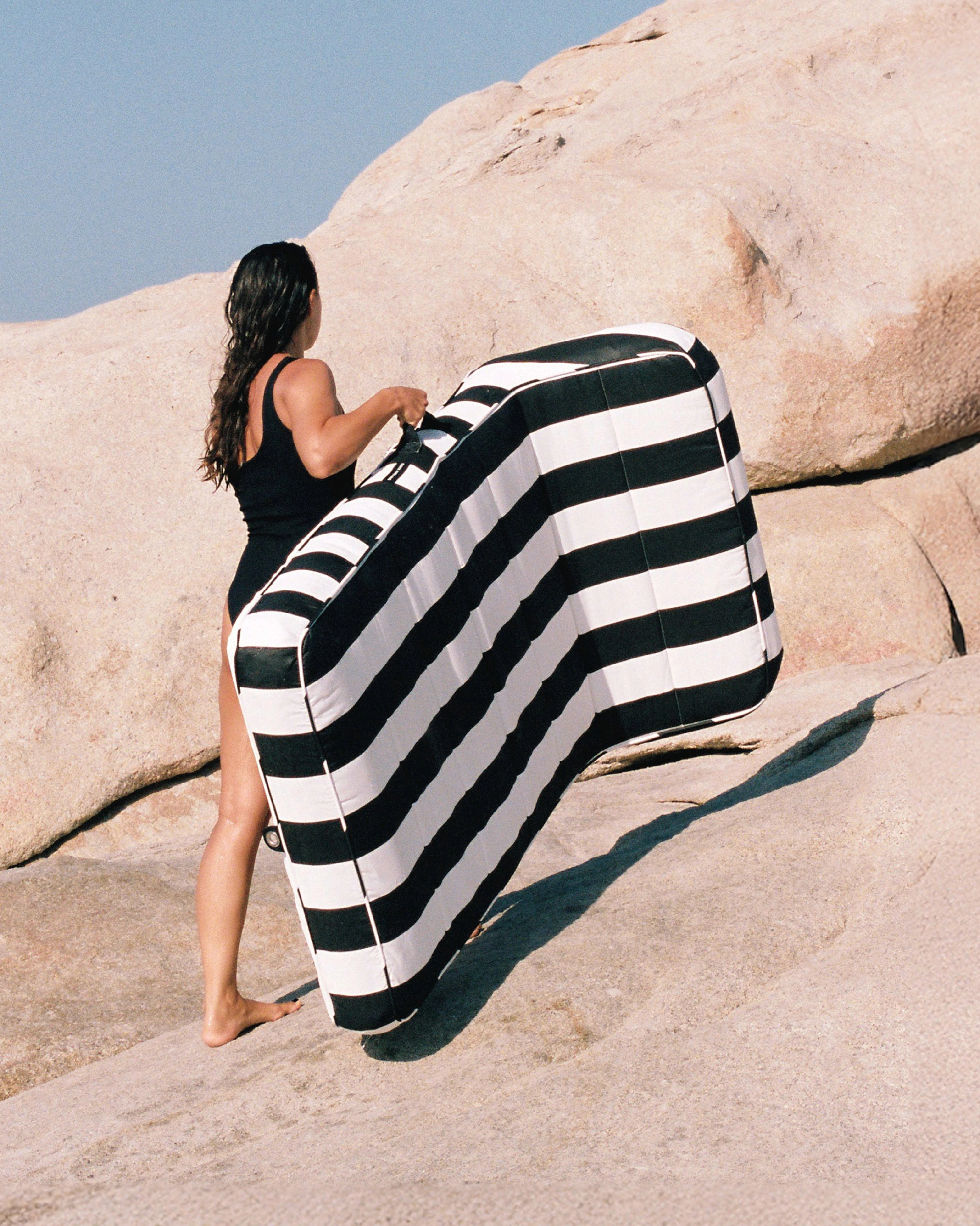Woman in a black swimsuit carrying a black and white striped luxury pool float across sunlit rocks — effortless portability meets bold design by Oliver James Lilos.